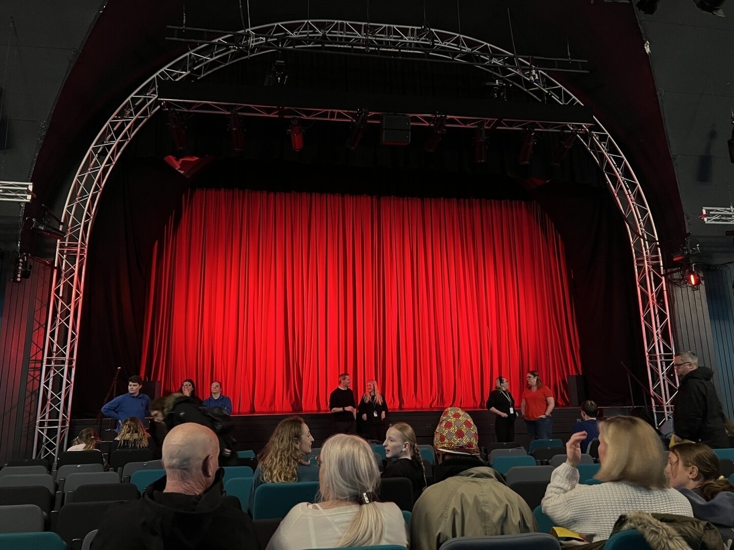 A theatre stage with a vibrant red curtain is viewed from the audience, where people are seated and interacting.