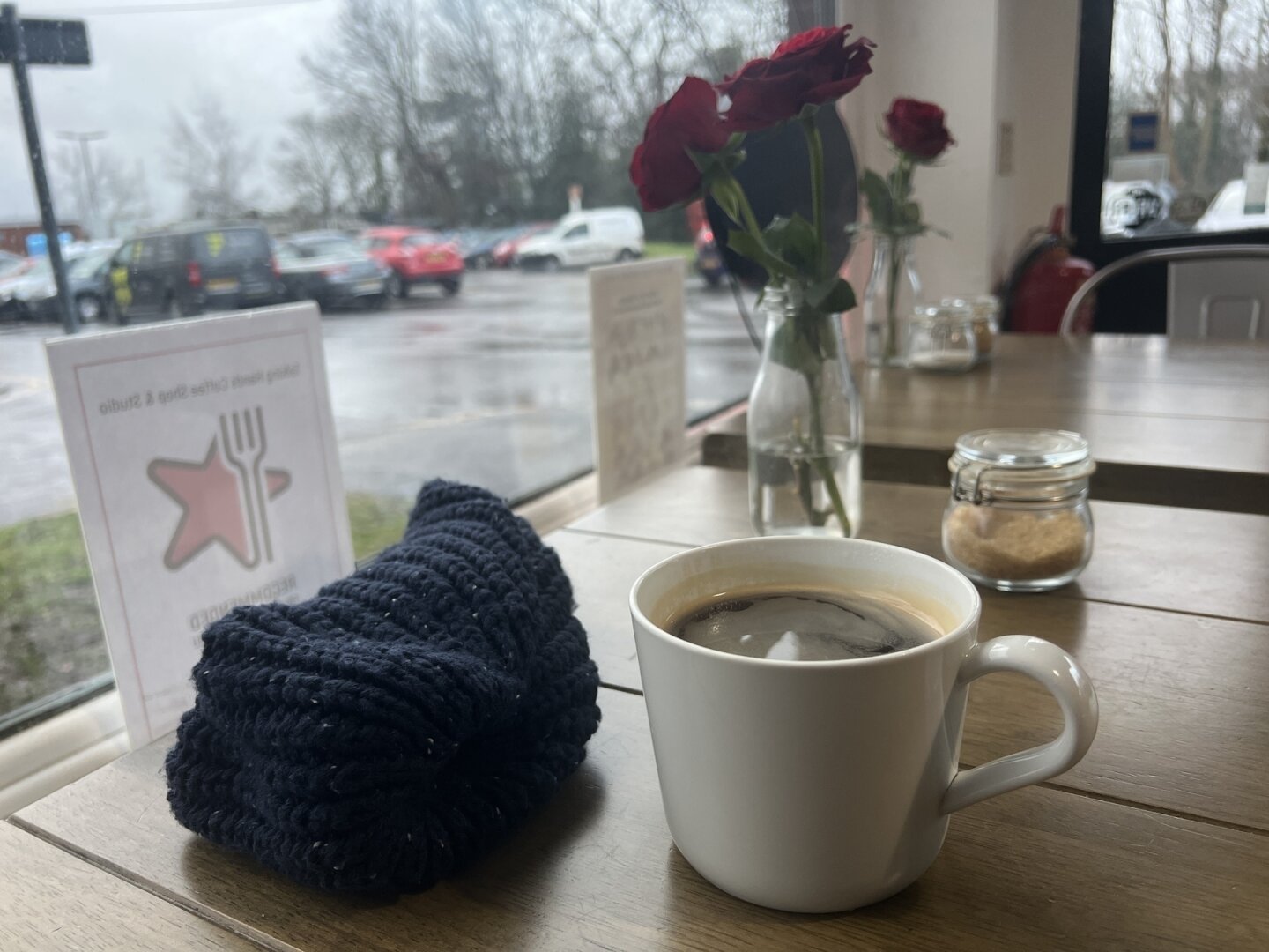 A cup of coffee, a knitted hat, and a vase with red flowers are arranged on a table by a window overlooking a station car park.