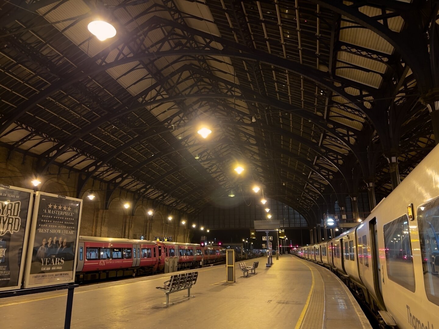 A train station platform is illuminated at night, featuring trains parked along the tracks under a large arched roof.