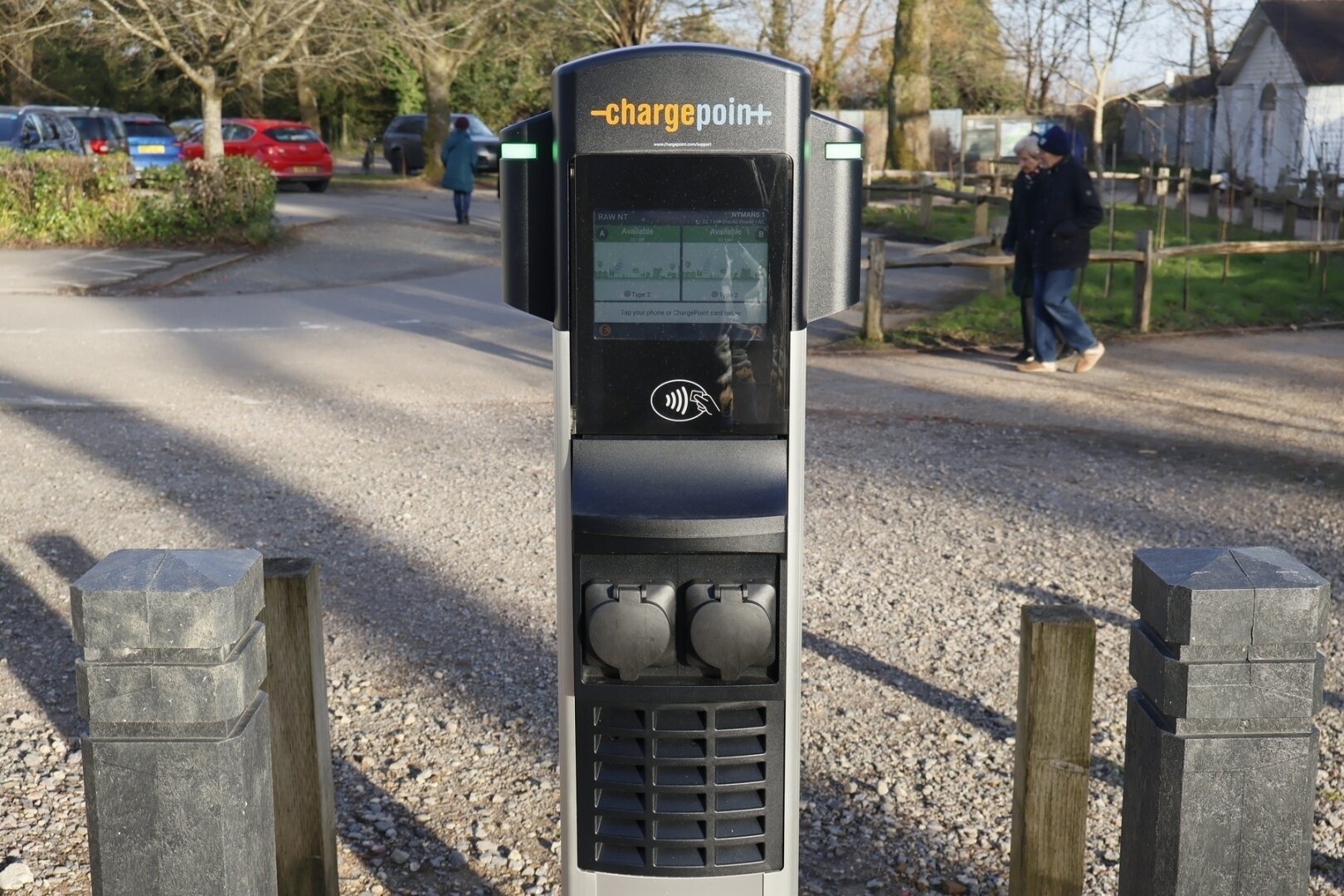 A charging station is situated along a pathway with parked cars and a person walking in the background.