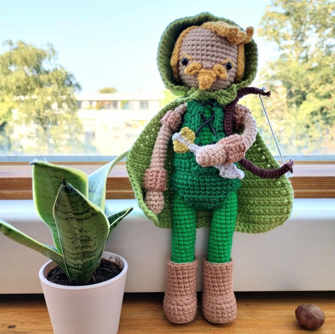 A crochet doll in green clothing and a green cape holding a bow and arrow.