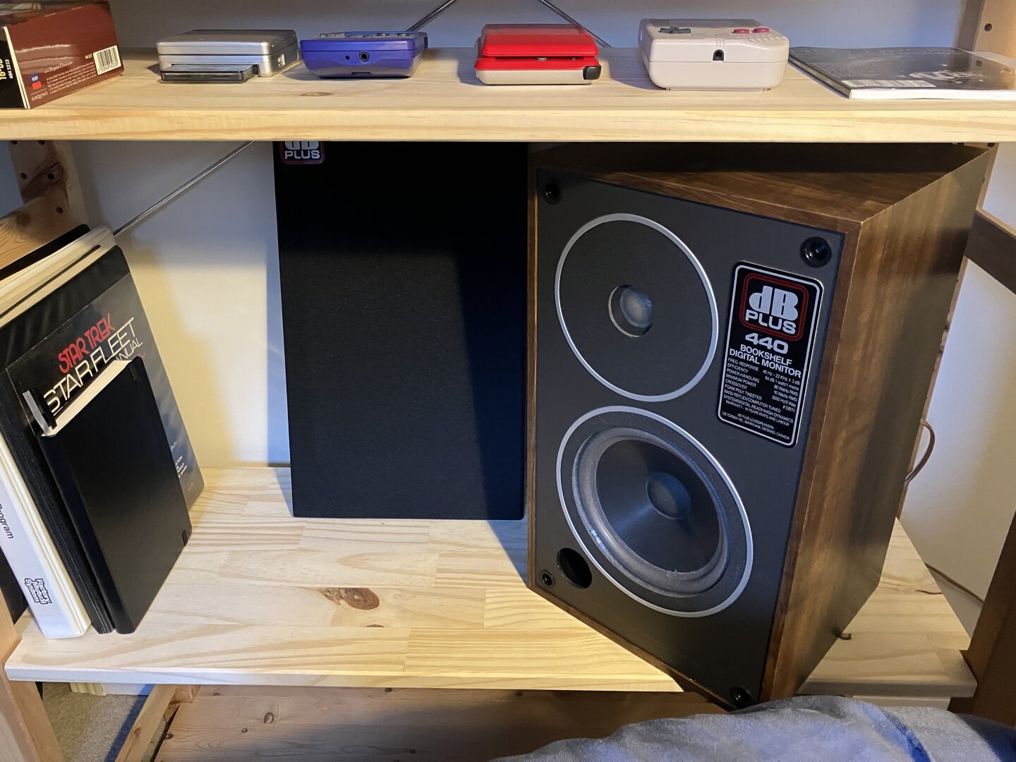 A vintage '80s era speaker on a shelf. The grill removed leaning beside it. Books to the left. Start Trek - Star Fleet Technical Manual amongst them.