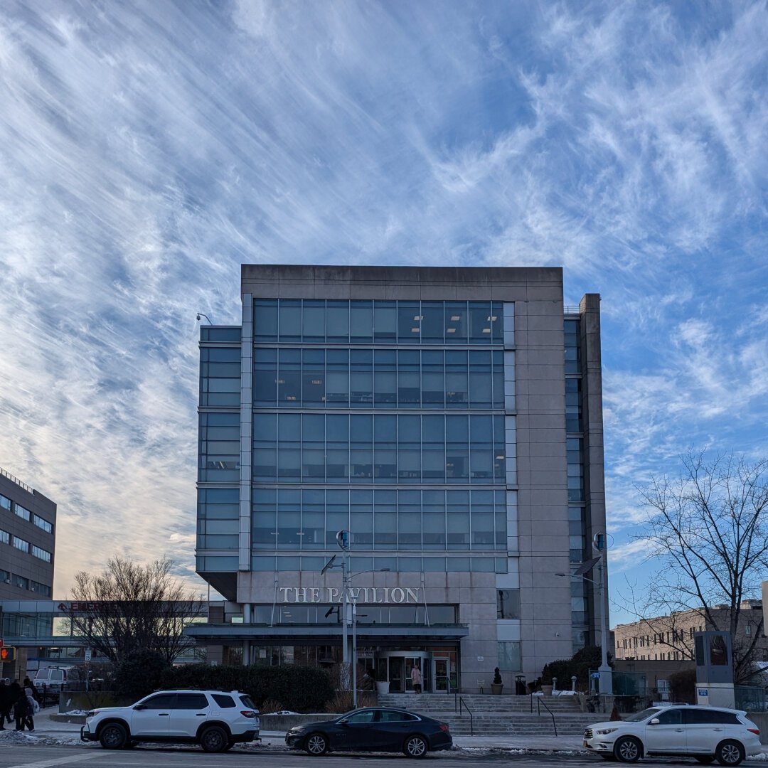 A six story, window lined building. The skyline behind it is a blue with streaky clouds.