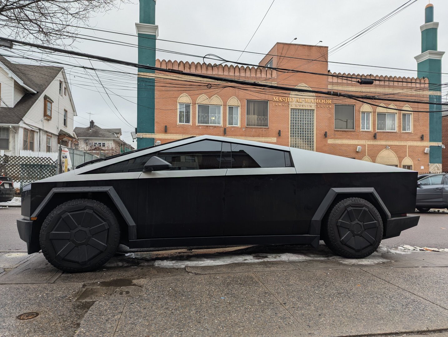 Black Cybertruck with tinted windows across the street from a red brick mosque with green minarets.