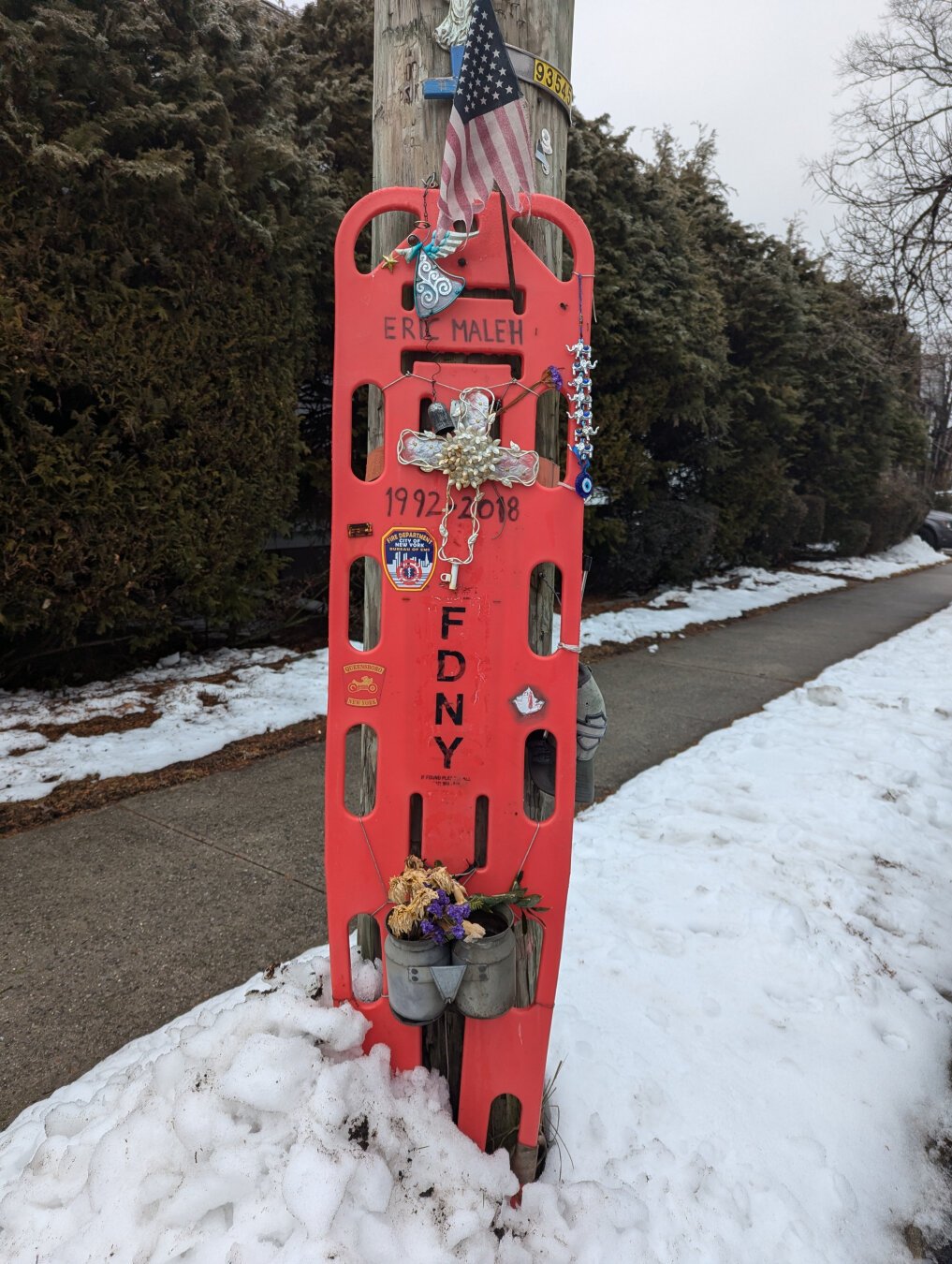 Plastic red-orange equipment branded with FDNY EMT logos with a cross and the name Eric Maleh, strapped to a power pole.