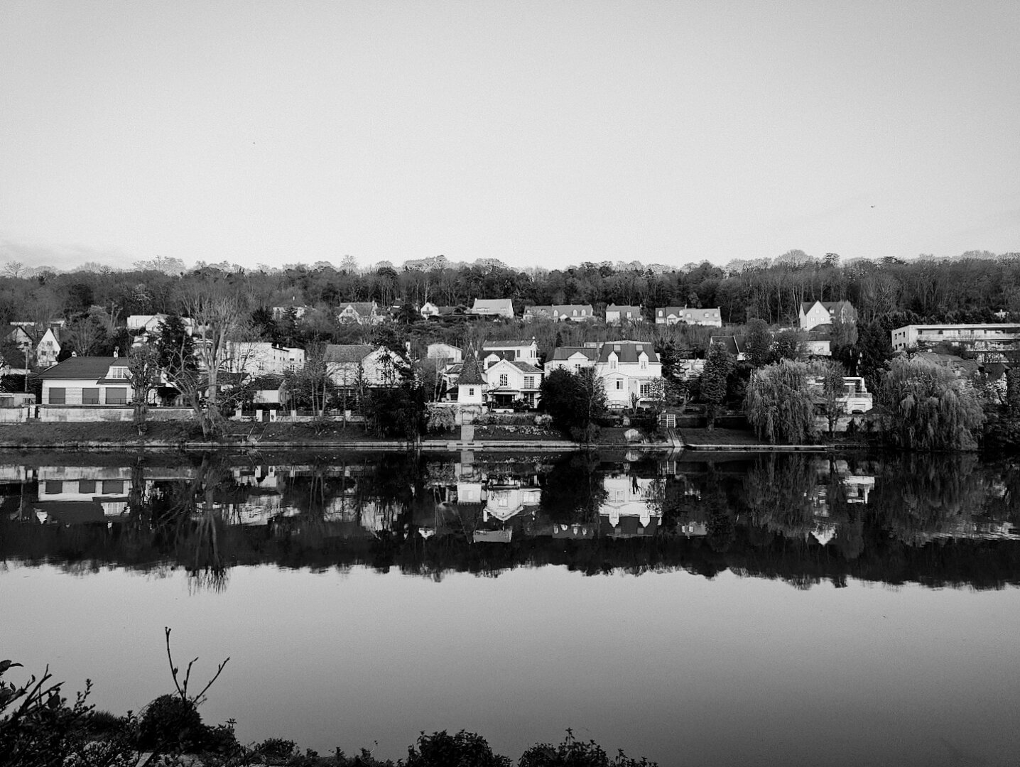 Une rangée de maisons borde un fleuve calme, leurs reflets nets dessinant un miroir parfait. La scène, en noir et blanc, évoque la tranquillité d’un village paisible niché entre eau et forêt. L’atmosphère est figée, presque hors du temps. La photo est prise à partir de la Promenade des Anglais à Saint-Maur-Des-Fossés.