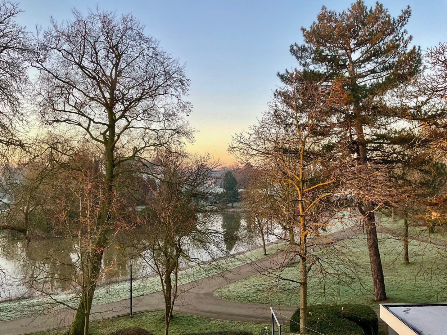 L’image montre un paysage matinal avec un parc bordé d’arbres partiellement dénudés, signe d’une saison froide. Le sol est légèrement givré et un plan d’eau reflète la lumière douce de l’aube. Le ciel clair et le ton doré du lever du soleil ajoutent une atmosphère paisible et hivernale.