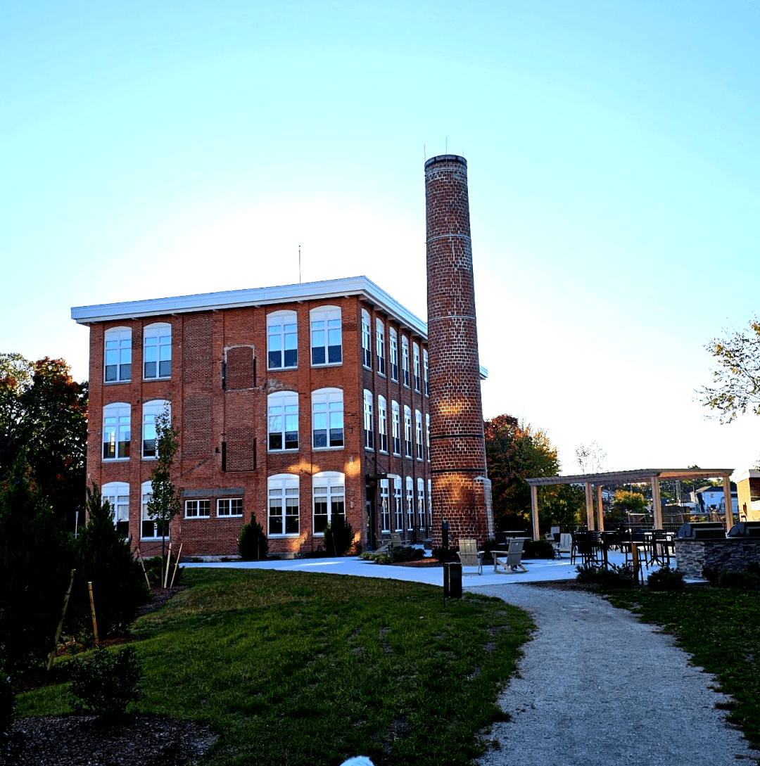 old brown brick mill building with white trimmed windows next to an old smokestack with a foot path leading to it