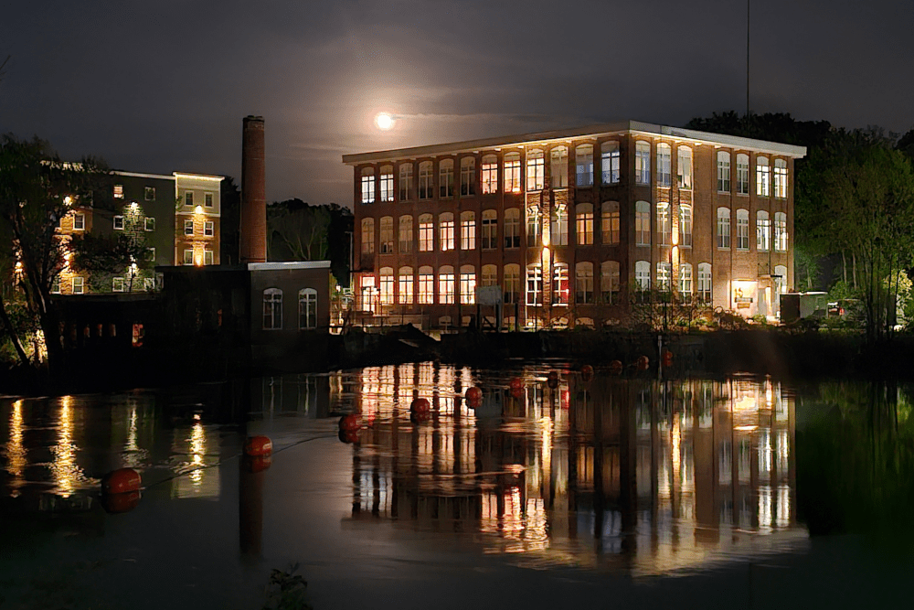 An old mill-style brick building with lights reflecting into the river creating a symmetrical image in the water. Colors of white, yellow, and red from the brick reflect off the water. The off-white moon sits behind a cloud above the building casting a hazy feel to the sky. Another building and smoke stack are seen to the left.