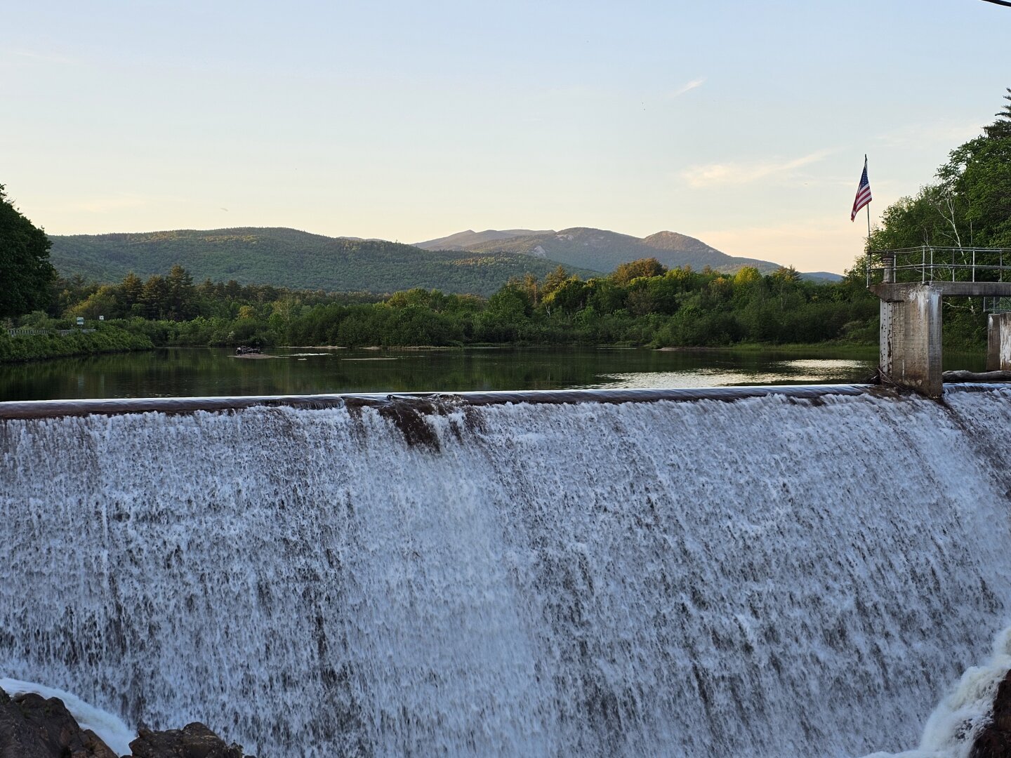 A dam with water flowing over with a pond in and mountains in the background