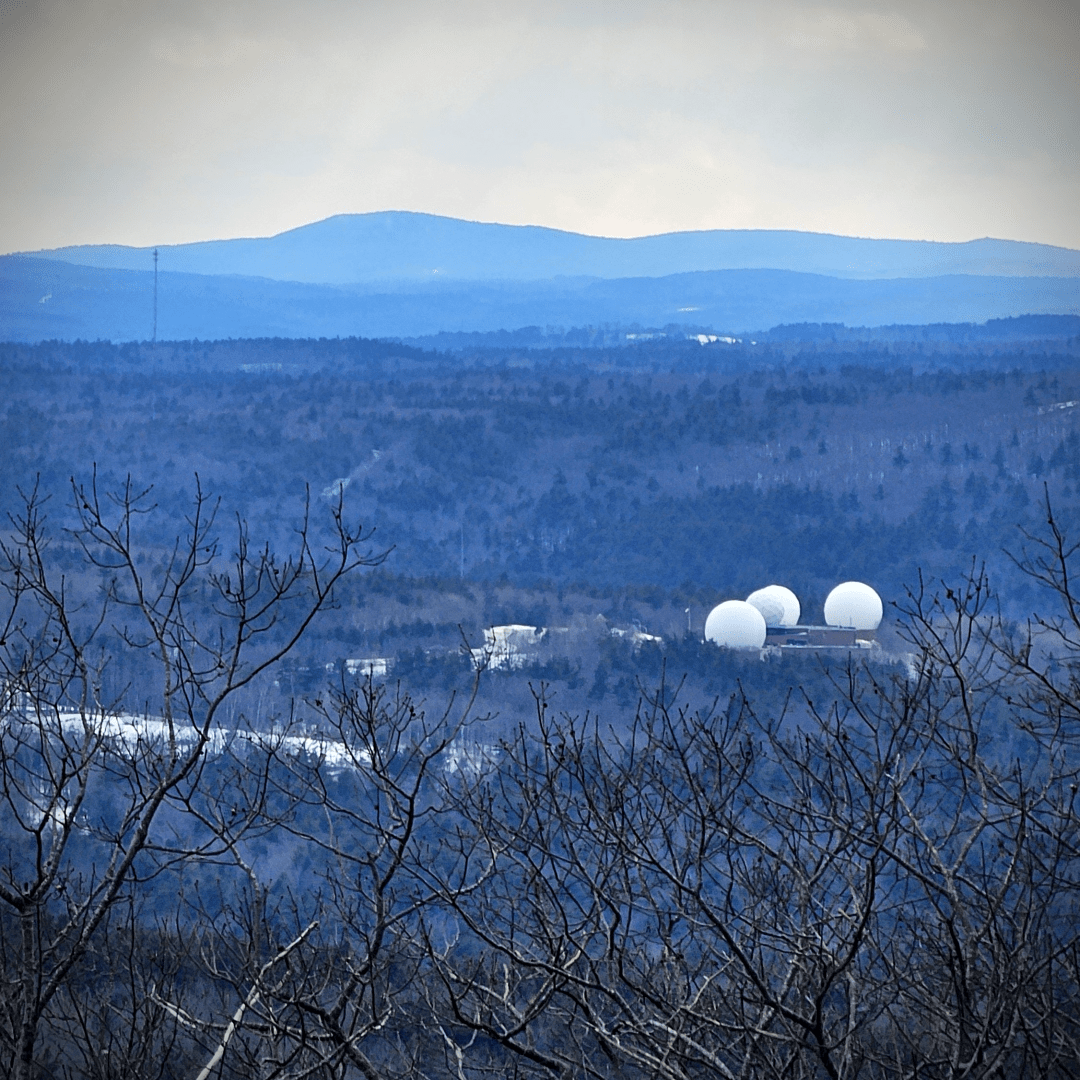 blue and gray landscape with trees in the foreground , white globes of a radar station in the ground and rolling hills in the distance