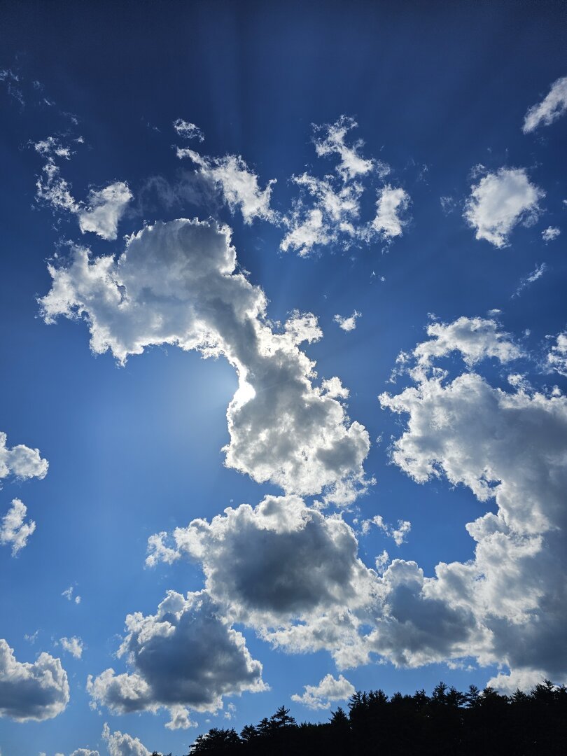 Sun rays behind large white puffy clouds and deep blue sky with tree line in the front