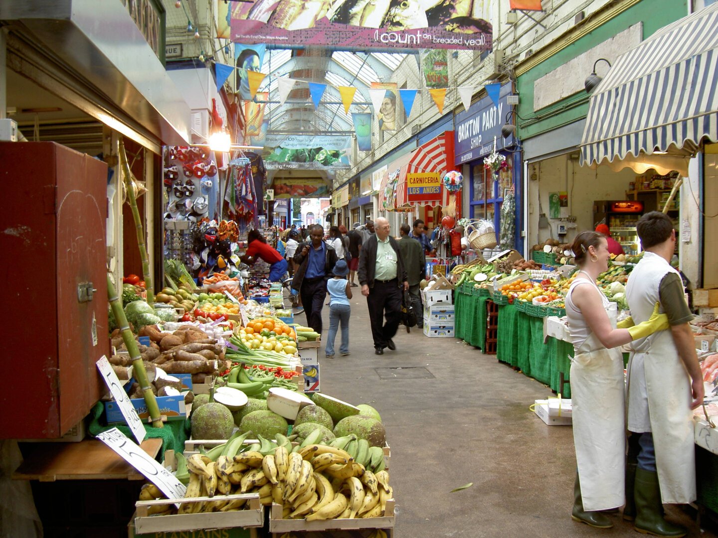 Indoor street with small shops on both sides. Vistors and workers. Foreground to the left is fruite shop with bananas and a lot more of fruits and vegatables follows further down in the foreground. To the right probably meat in the shop in foreground, difficult to tell. Also in the right foreground two woekers, a female looking worker with yellow gloves holding her hands from back holding a male looking worker standing in front of in direction to the meat shop. They have rubber boots and long platic aprons suitable for meat or fish work. Then on right side follows fruit and vegetable shops. And several other shops on both sides, not only eatable goods. Some glass in the ceiling and various decoration.