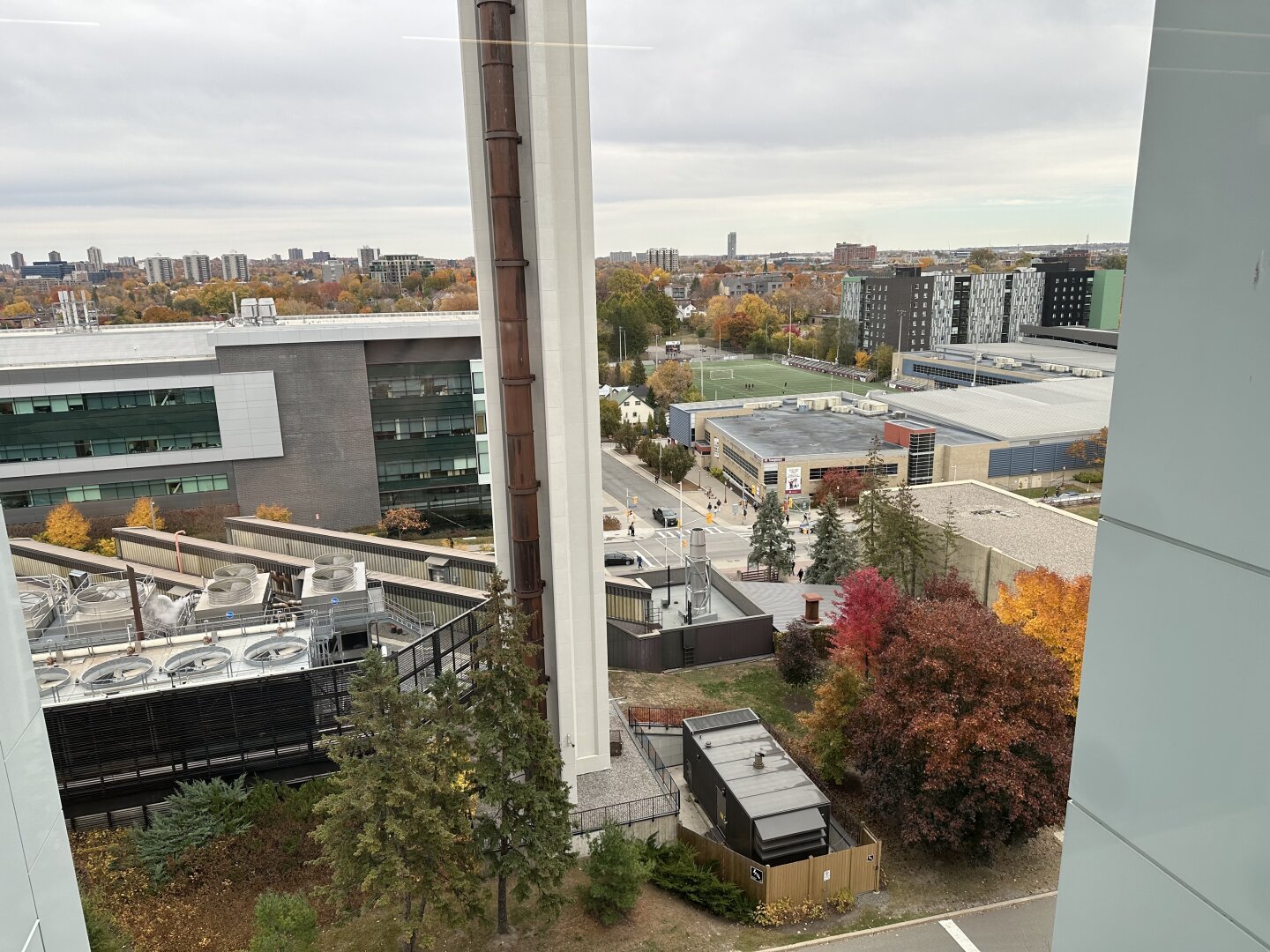 Cluttered view of urban area from a 6th floor level window. Some trees but mostly concrete structures. There is a green pitch in the distance.