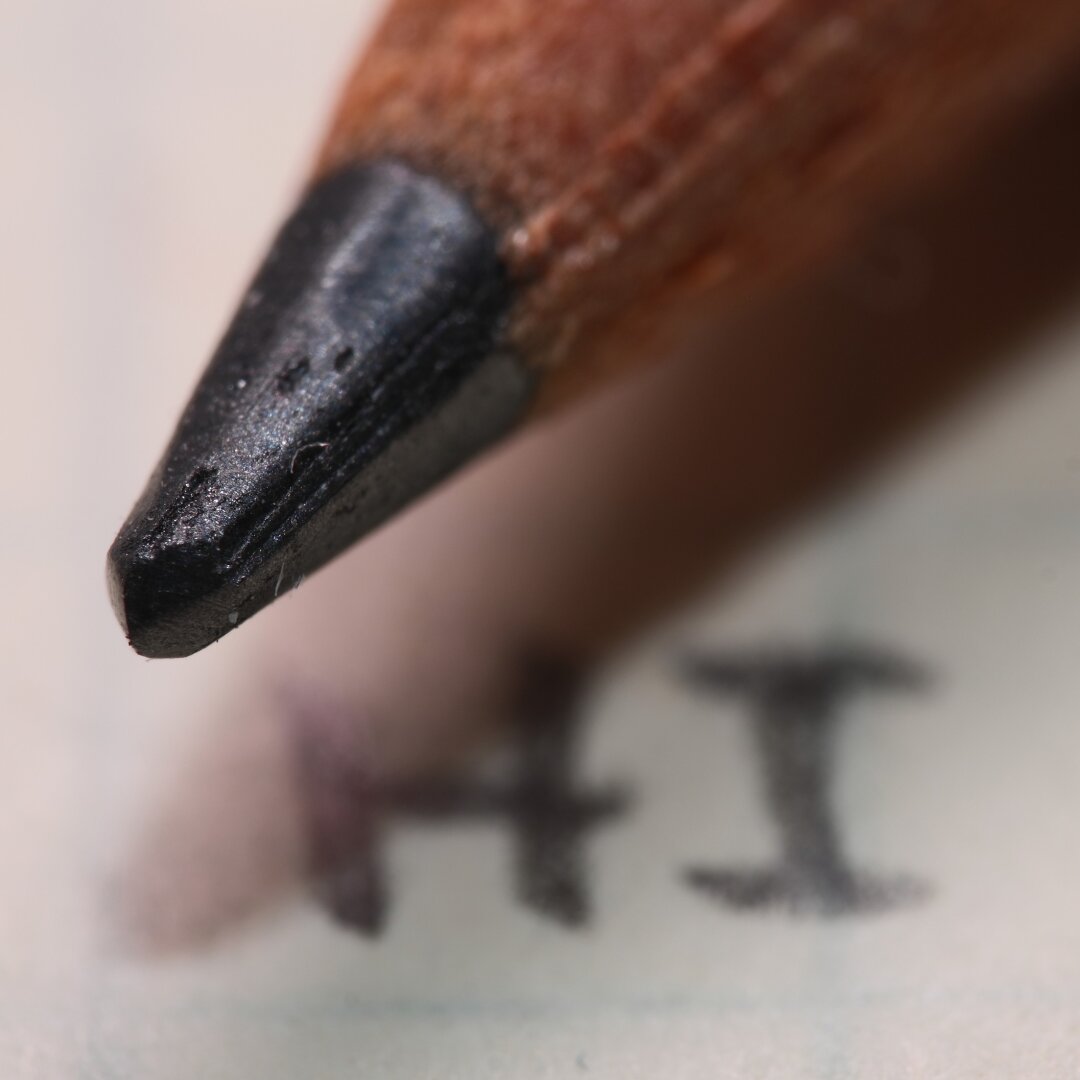 A macro photo of the tip of a pencil, it is sitting on a paper that has the word HI written on it.