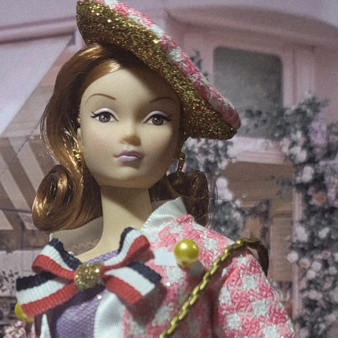 Close up photo of a Susie doll by R&D, wearing a pink checkered Chanel-style jacket, over a purple top with a big bow with blue, white and red stripe. She also wears a beret, with the top fabric similar to her jacket. The bottom part of her beret is gold that matches the chain of her Chanel-style bag.