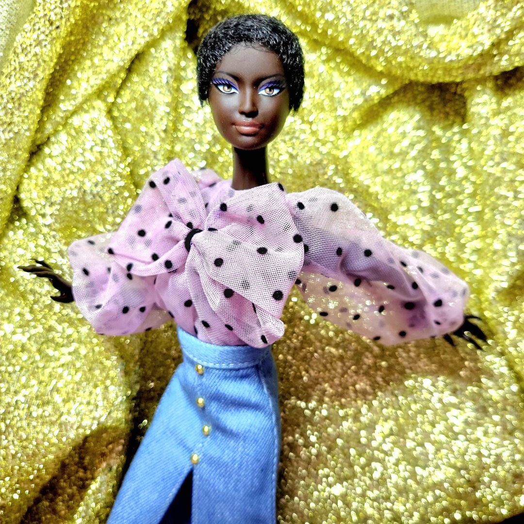 A dark-skinned Barbie doll with short curly hair wearing a sheer long-sleeved lavender with black polka dots blouse with a big bow on the front and denim skirt.