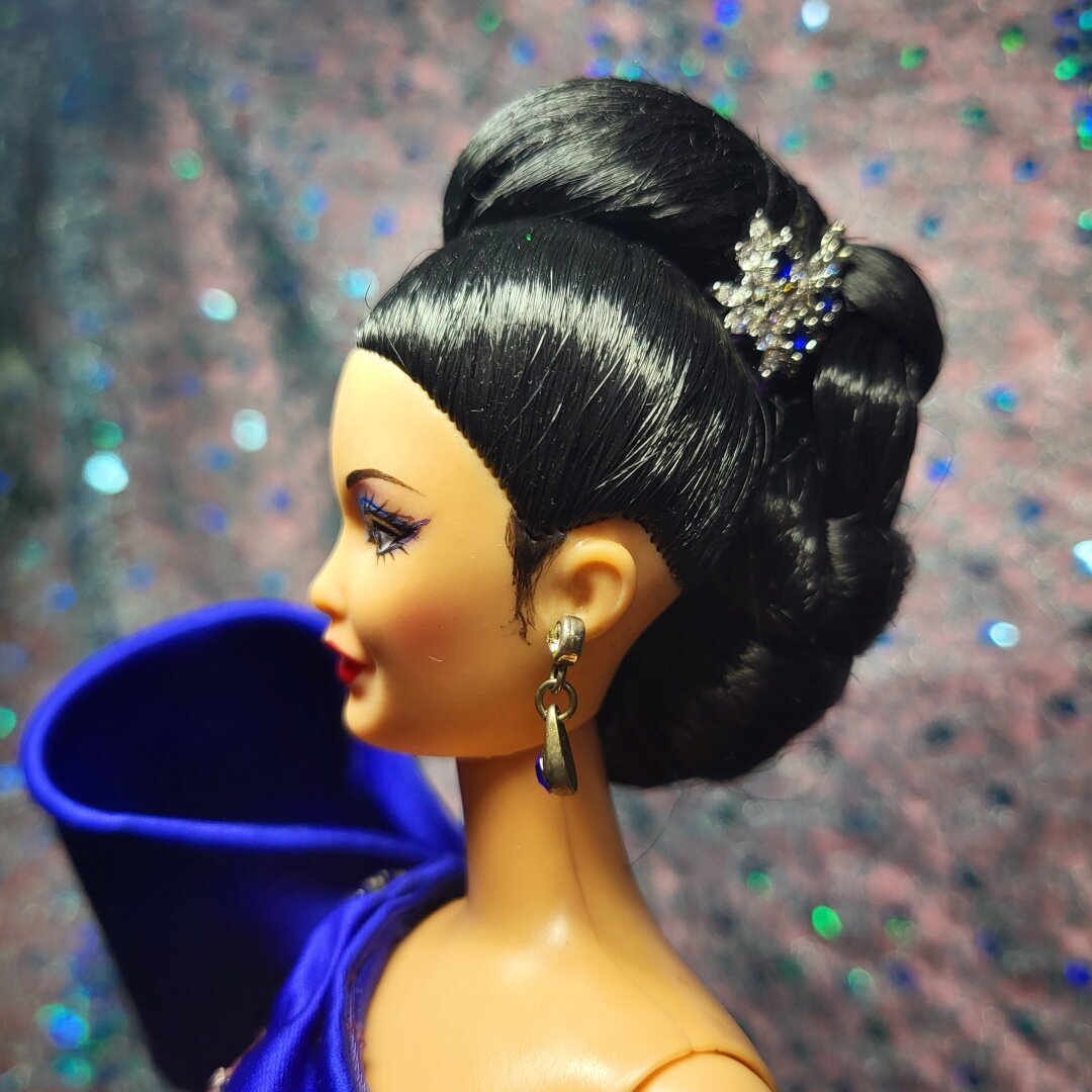 Barbie doll with Asian features, long black hair braided into an asymmetrical bun adorned with human earrings having clear, and blue crystals. The blue crystals on the hair matches her blue eyeshadow, blue gown, and her blue doll dangling crystal earring.