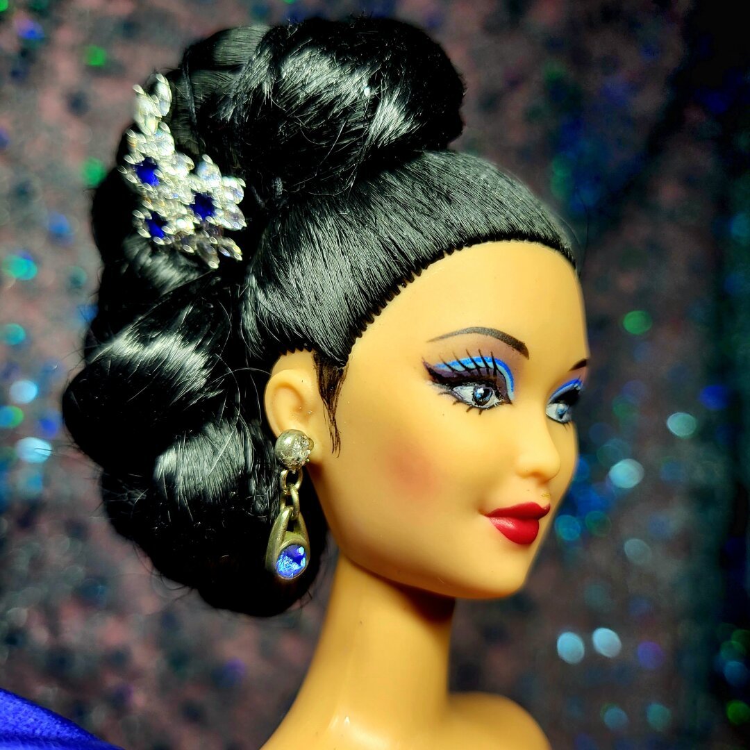 Barbie doll with Asian features, long black hair braided into an asymmetrical bun adorned with human earrings having clear, and blue crystals. The blue crystals on the hair matches her blue eyeshadow and her blue doll dangling crystal earring.