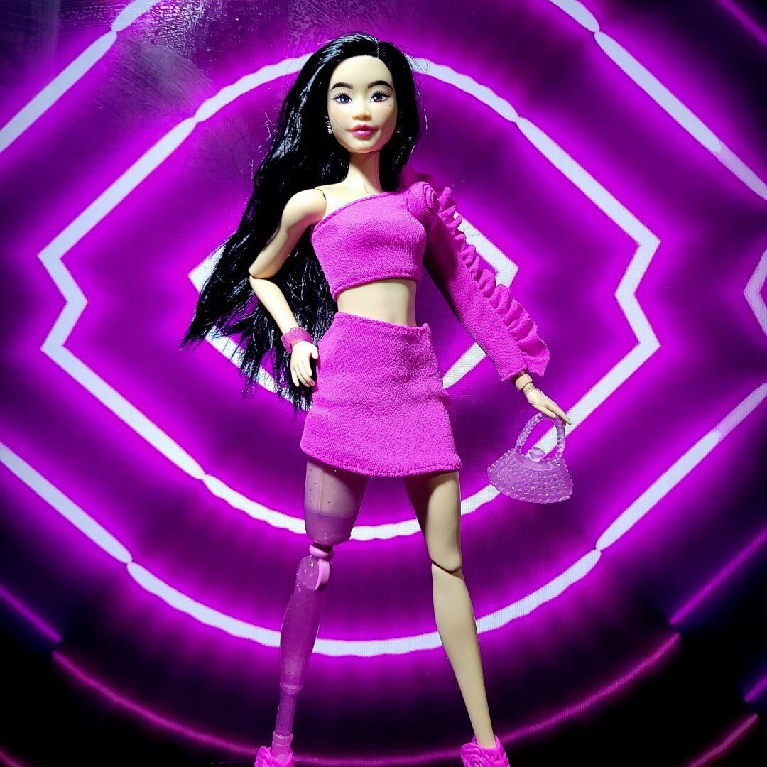 Amputee Barbie doll with a pink prosthetic leg wearing a pink asymmetrical blouse with ruffled sleeve, and a matching mini skirt.