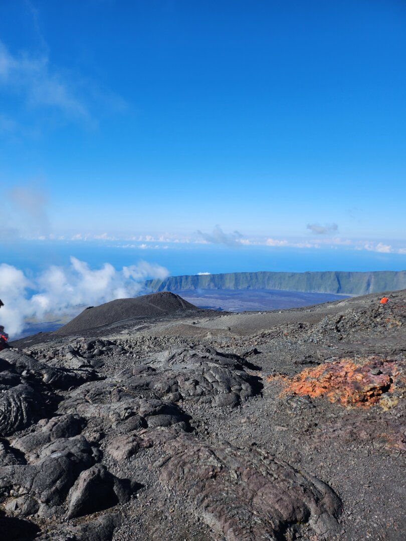 A view of a small sloping crater with cooled lava in the foreground, the ramparts surrounding the volcano and some clouds in the background and a blue sky above the sea in the distance.