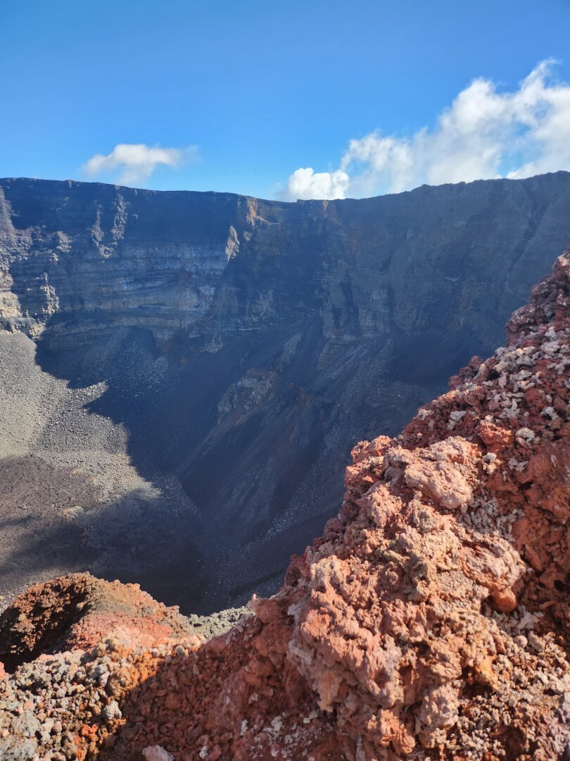 A view of the interior of the Dolomieu crater from the summit of the volcano, with orange volcanic rocks visible in the foreground.