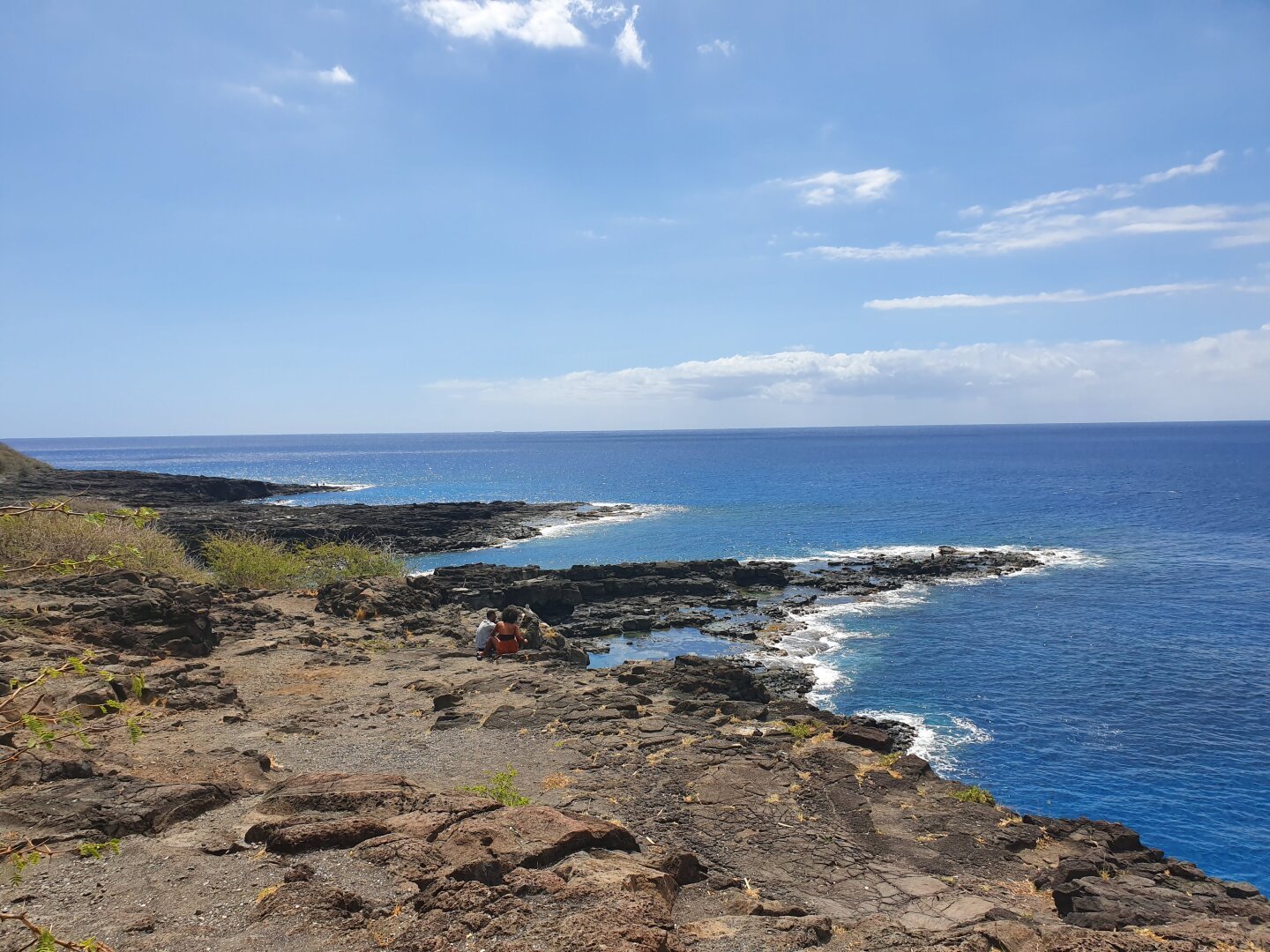 un petit bassin naturel au bord de la mer et des jetées naturelles formés par de la roche volcanique, un couplé est assis, quelques plantes sont visibles