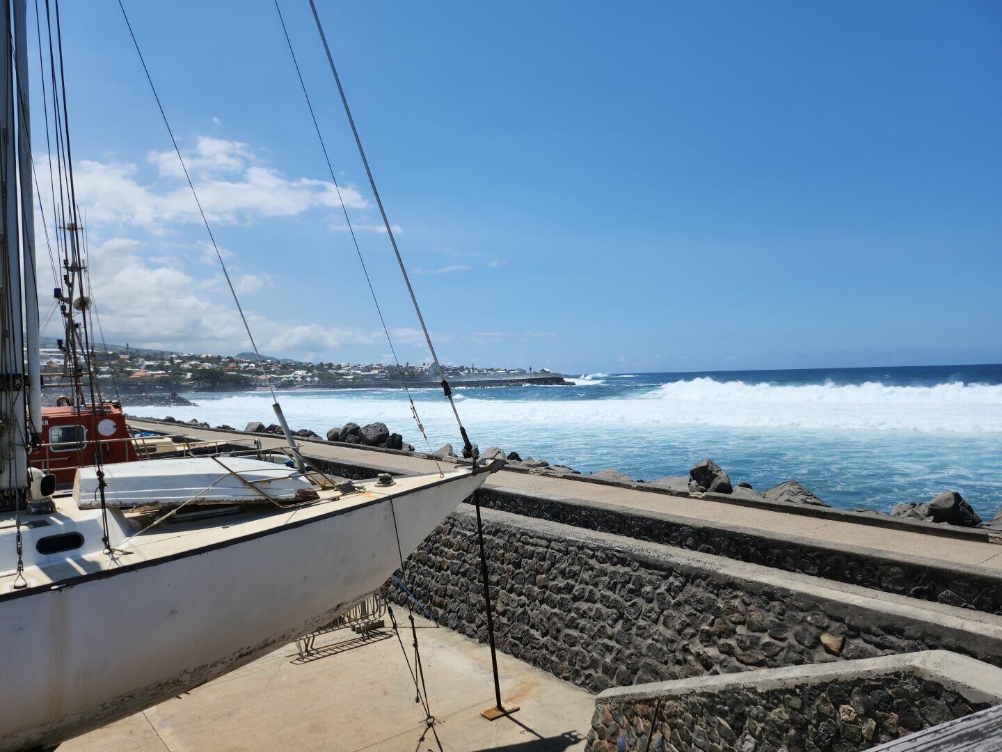 Photo taken from a harbour, the sky is blue, two boats can be seen in a parking lot on the left in the foreground, a slightly rough sea in the background and inhabited areas in the background on the left.