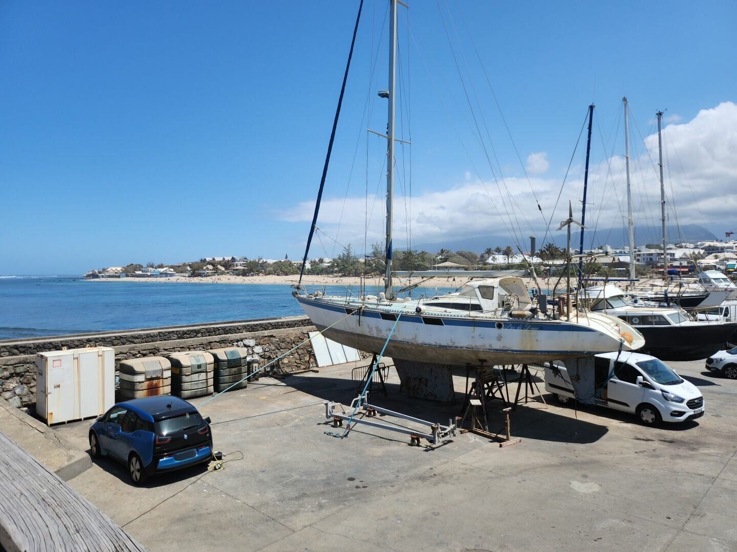 Photo taken from a harbour, the sky is blue with a cloudy mass, several boats and cars can be seen in a parking lot in the foreground, you can see the beach and the sea in the background