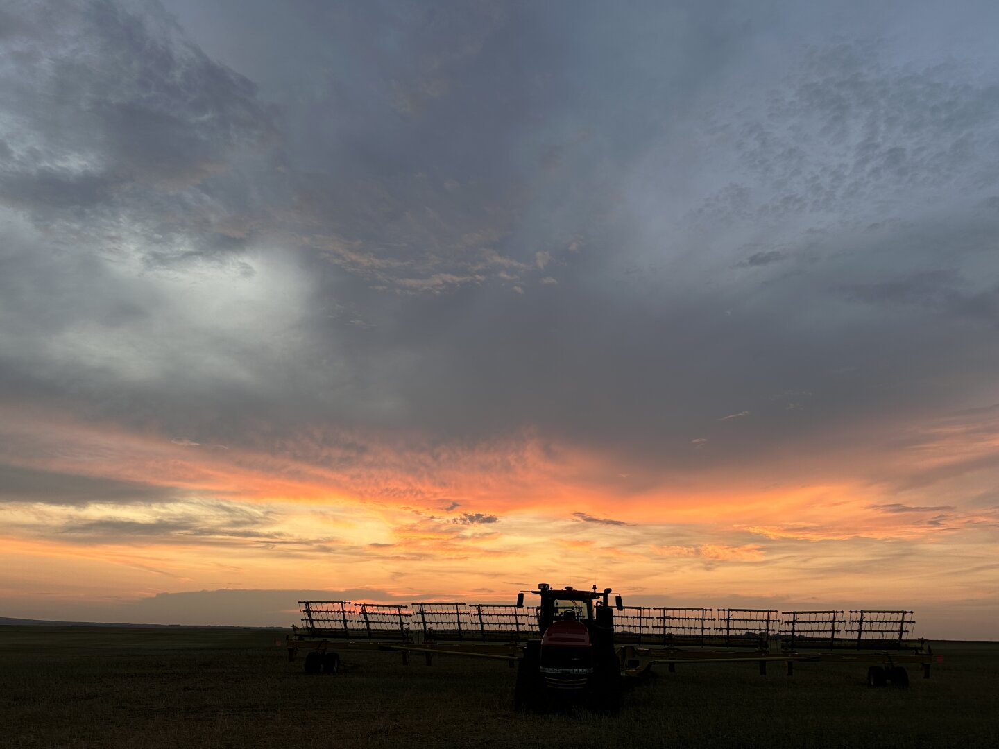 Farm tractor and machinery on dark horizon below a brilliant vibrant sunset set in heaven clouds above.