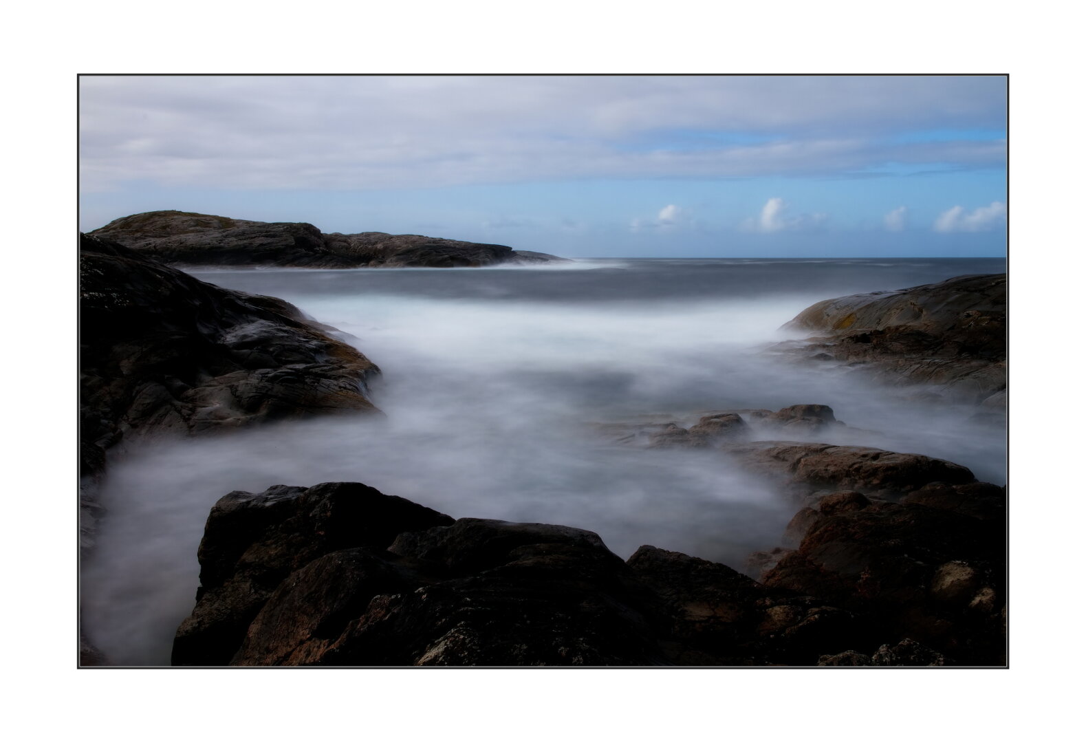 Long exposure photo. Waves between the rocks. Under the partially cloudy sky. Herlevaer, Norway