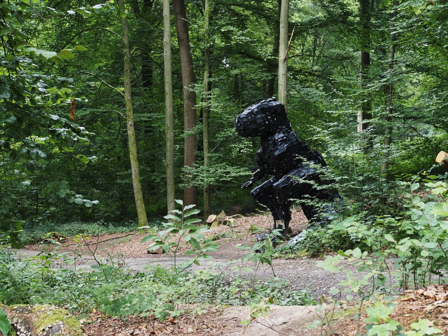A dinosaur in the woods. It seems to be a T-Rex made from a wooden structure covered with old video tapes.