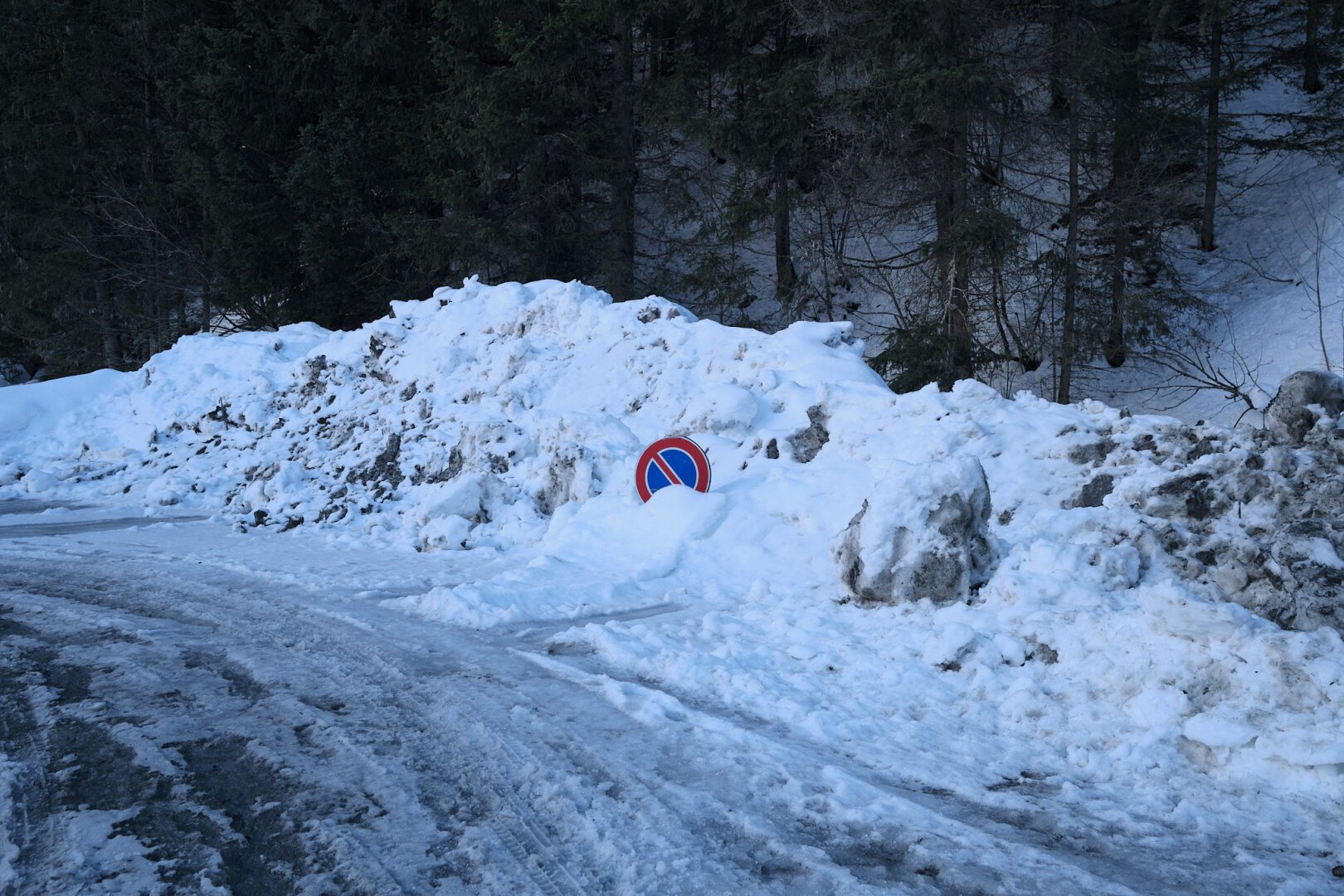 A red and blue forbidden stop sign is barely visible in a mountain of snow, on the roadside of a slippery snowy road. The image has a blue hue to it