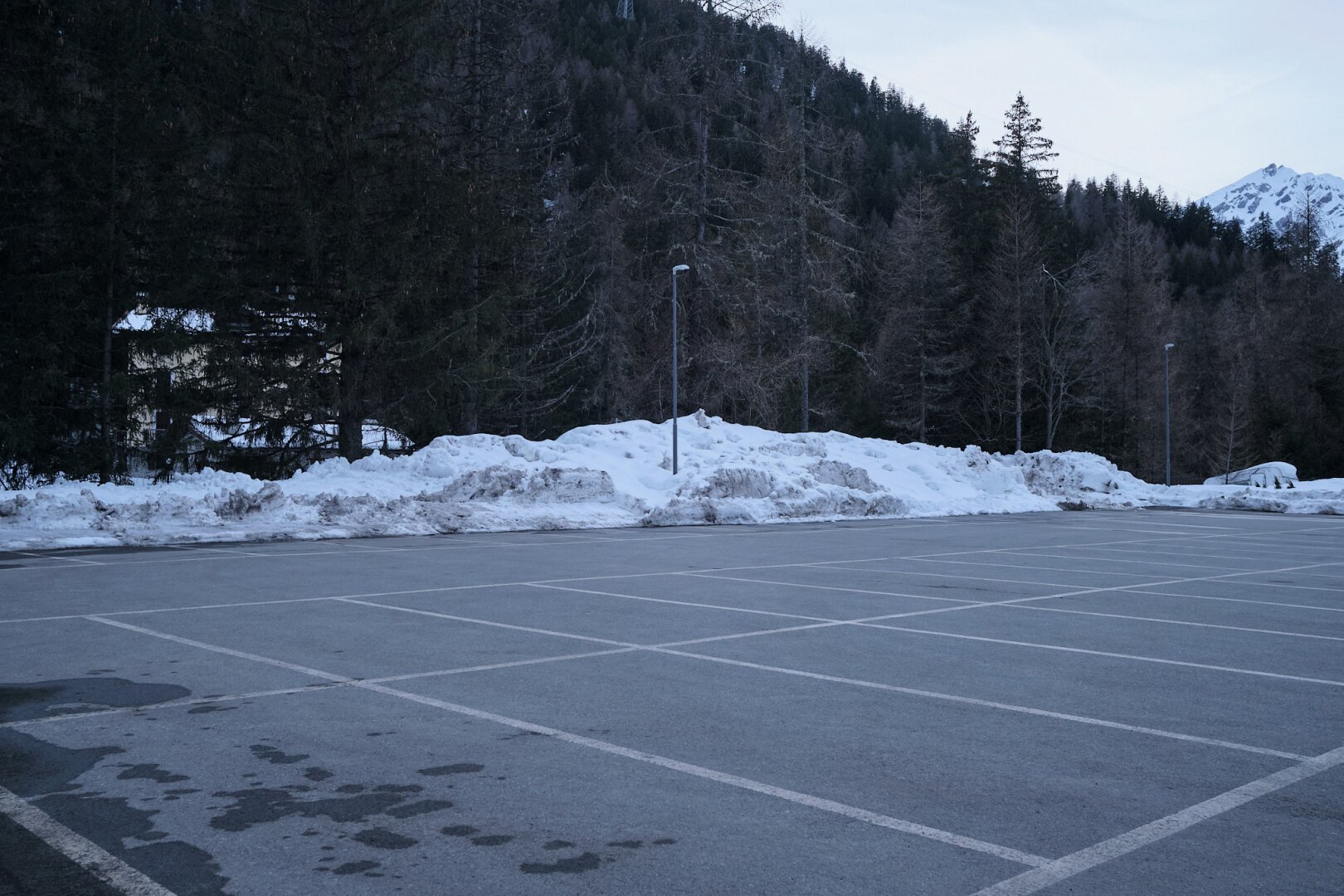 A deserted parking lot on the Alps, with snow shoveled all around its sides. There's a solitary lamp post sprouting from the snow.