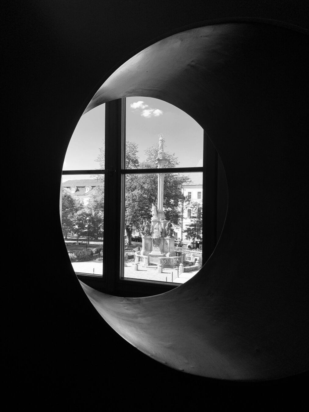 View through a circular window framing a historic monument in a town square, with trees and buildings in the background, captured in black and white.