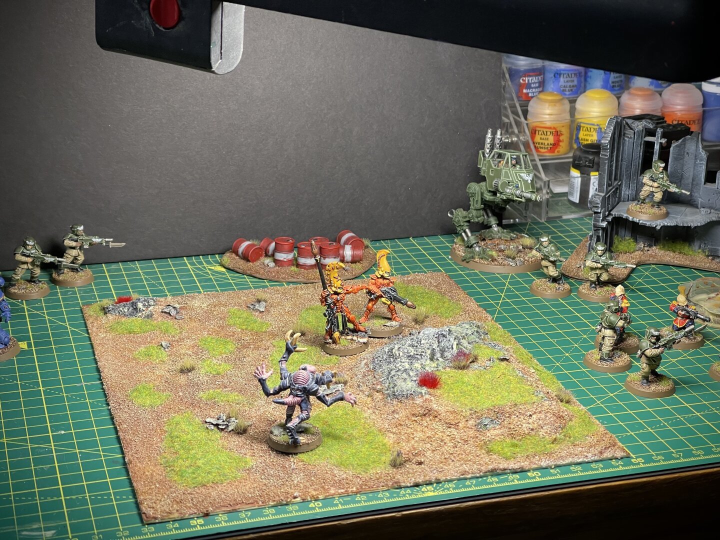 Mini wargaming board on painting desk, with some small hills, dirt, grass and tiny shrubs. An assortment of painted miniatures laid out on and around it