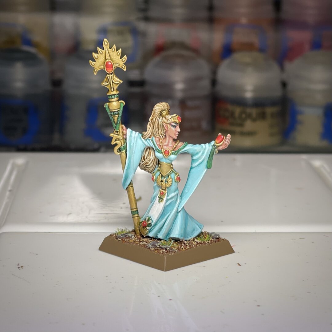 Painted Alarielle the Everqueen, classic 90s Warhammer. Flowing sky blue robes, golden hair, ornate staff. Adorned with gemstones, painted base. On painting desk