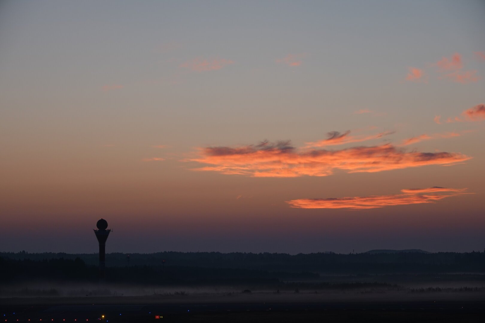 Bright orange Cirrus clouds on a morning sky. The sun has yet to rise. The sky is pretty dark on the horizon due to slightly reduced visibility. A ground traffic radar tower is shilouetted against the sky on the left. Shallow fog covers most of the ground. Taxiiway lights can be seen on the very bottom of the image.