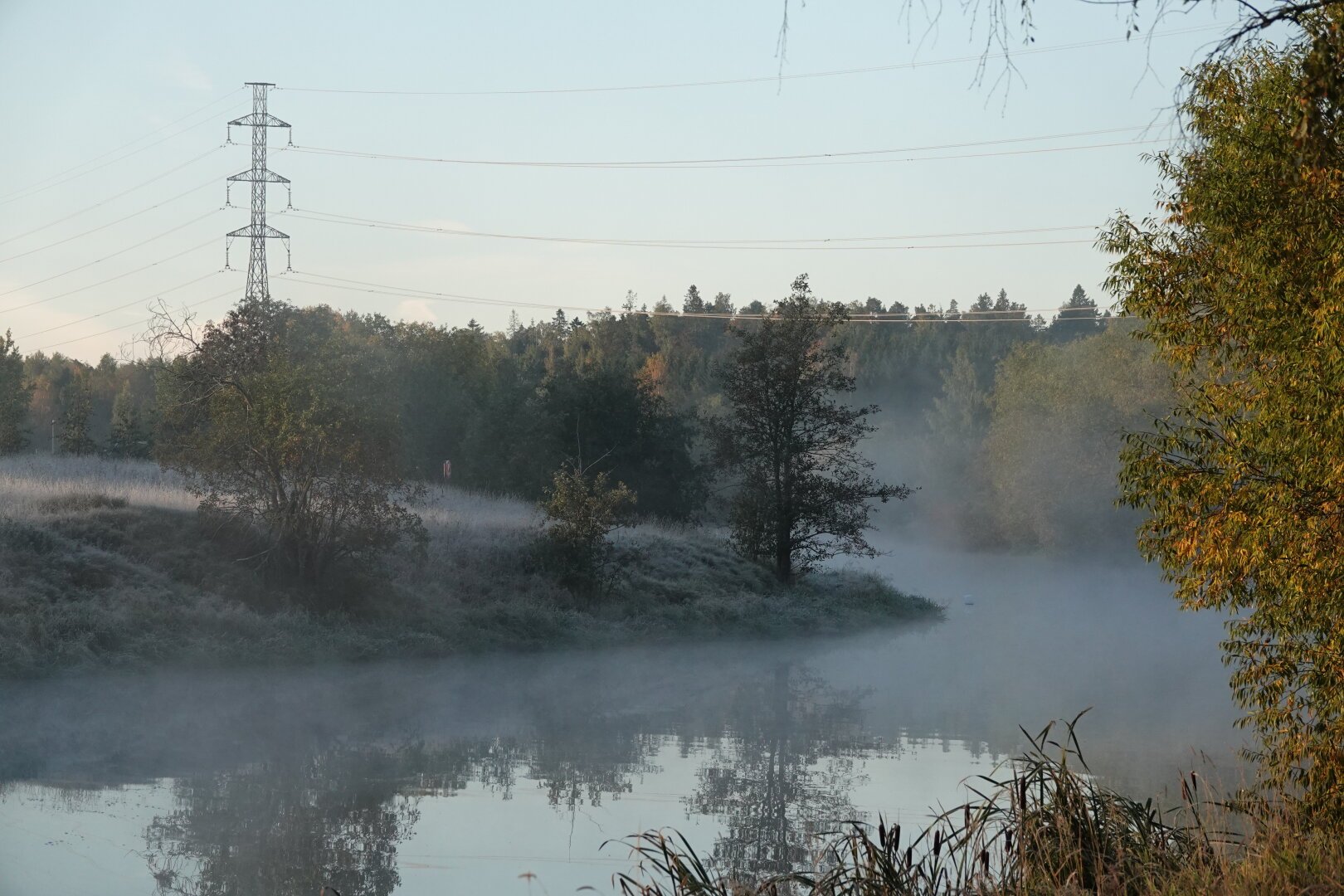 A misty and mirror calm river on a clear morning. A field on the other side of the river is white with hoar frost. Some trees have yellow leaves while others are still more or less green. A power line can be seen on the background.