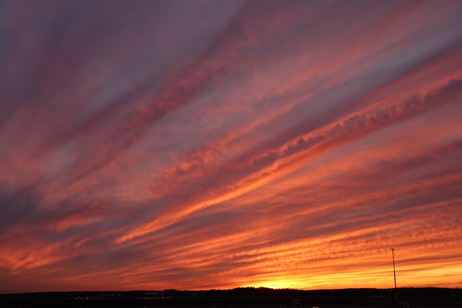 Orange sunset clouds. The sky is covered in clouds illuminated from below by the setting sun. Whispy linear structures give rise to brighter highlights and darker shadows where the illuminated clouds are more opaque. A pale gray blue sky glows through the thinner parts of the clouds.