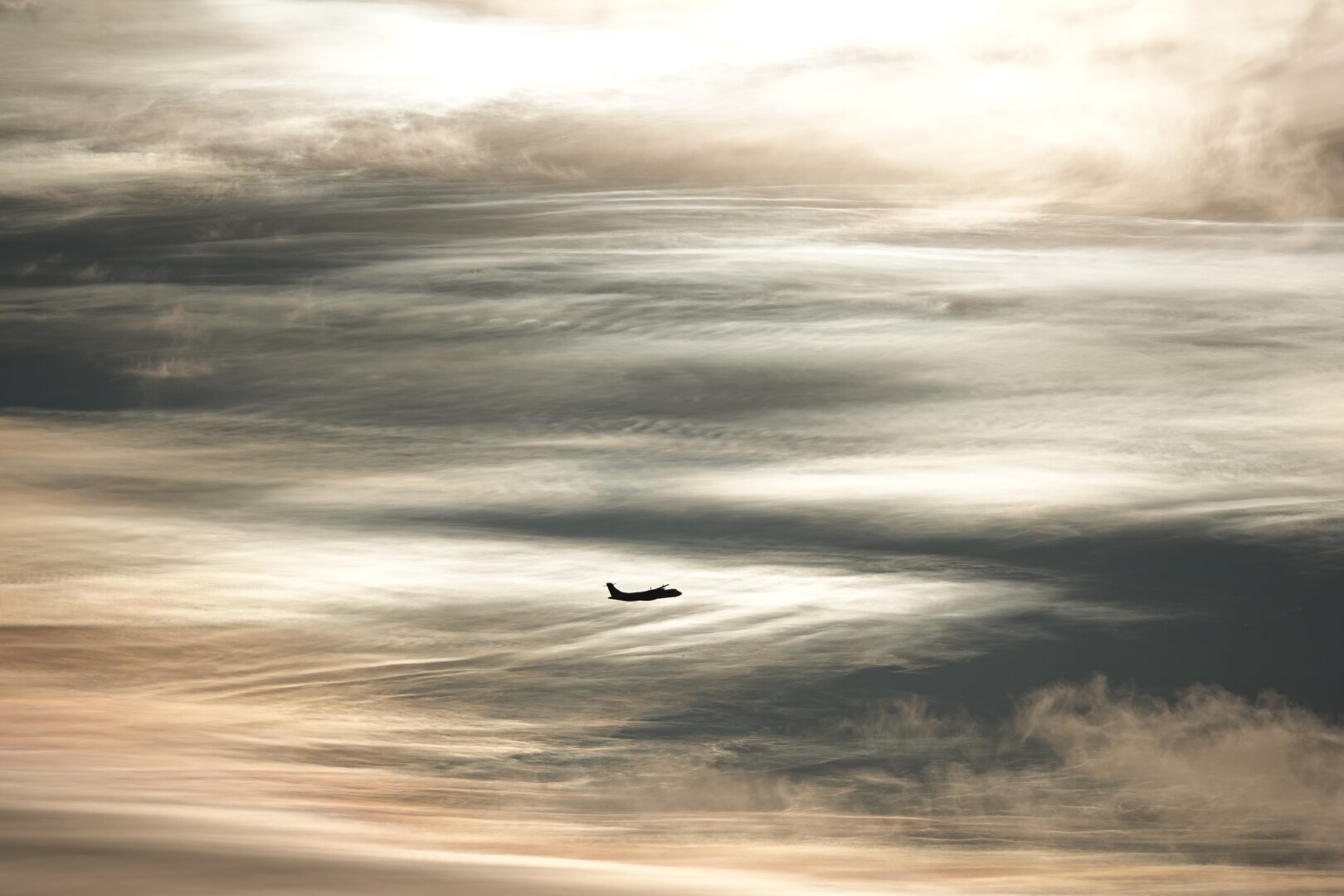 Slightly below the centre of the image there is the small black shilouette of a departing twin propeller aircraft, heading to the right, slightly away from the camera and with its nose tilted a bit upwards.
Behind the aircraft is a strikingly white mass of delicate cloud, which continues to the left and connects there to a larger sheet of varying density which spreads around from there to cover most of the image. The clouds are rich in detail, with gentle ridges and ripples here and there. Some of the ripples appear to exhibit the characteristic evenly spaced hooks of Kelvin-Helmholtz waves, also known in this context as fluctus. The clear sky behind the clouds is dark in comparison. At the edges of the image the clouds gain a slightly warmer tint as the diffracted sunlight seperates into different wavelengths at larger angles from the sun.
The image is subtly framed at the top and the lower right by lower clouds of similar colour, but fuzzier, less detailed texture.