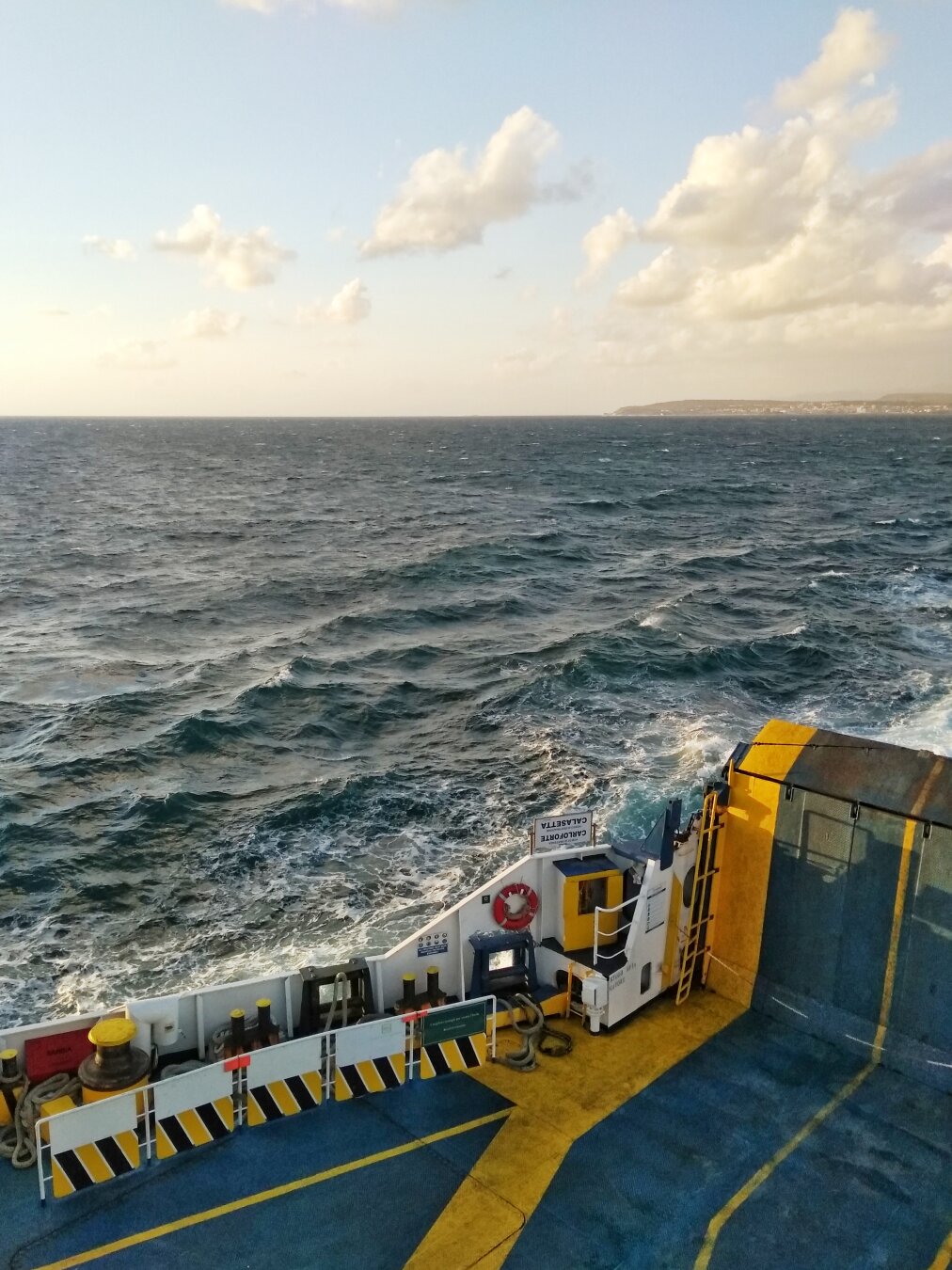 A photograph taken from the deck of a ferry, looking out over the dark blue Mediterranean Sea towards a distant shoreline under a partly cloudy sky. The foreground shows the yellow-marked deck of the ship and a safety ramp.