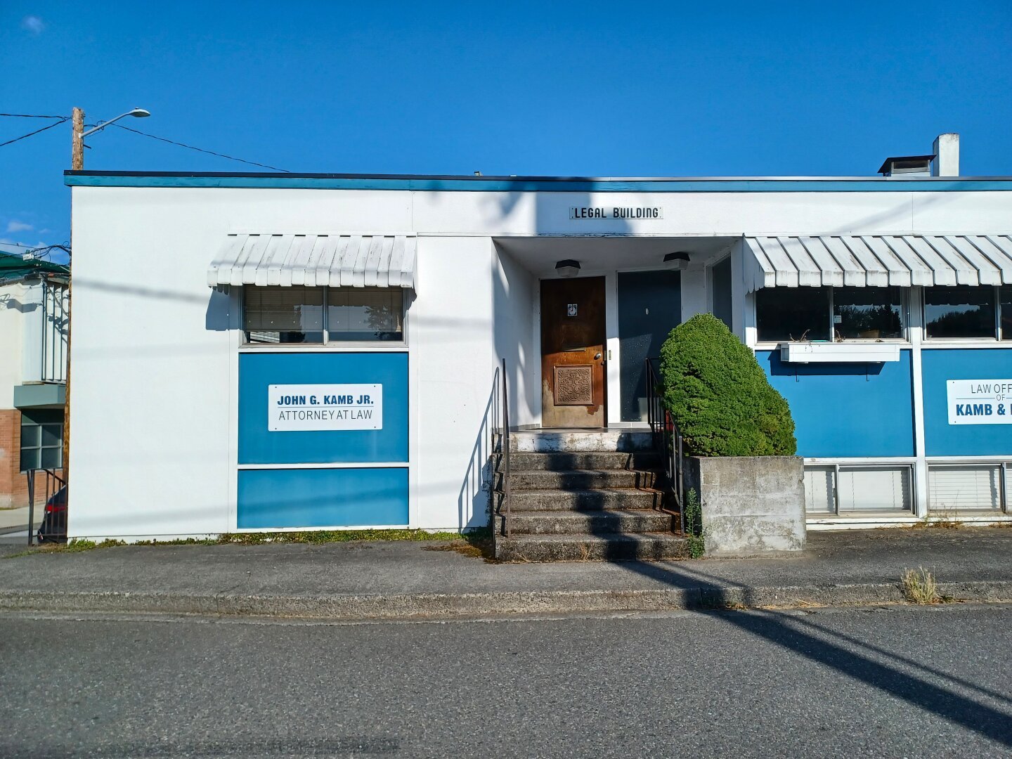 White and blue flat-roofed, midcentury concrete building with white vintage metal awnings. Building is white with blue trim. A small sign over the front door says 