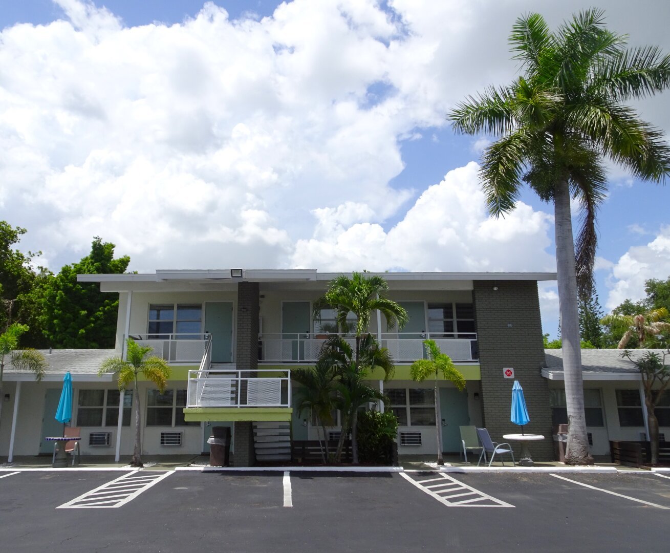 Exterior of a midcentury modern motel with a palm tree and concrete picnic tables out front. The two-story building have exterior stairs painted chartreuse green, white facade, and dark grey painted brick accents. The picnic tables feature bright turquoise umbrellas. Blue sky and puffy white clouds in the background.