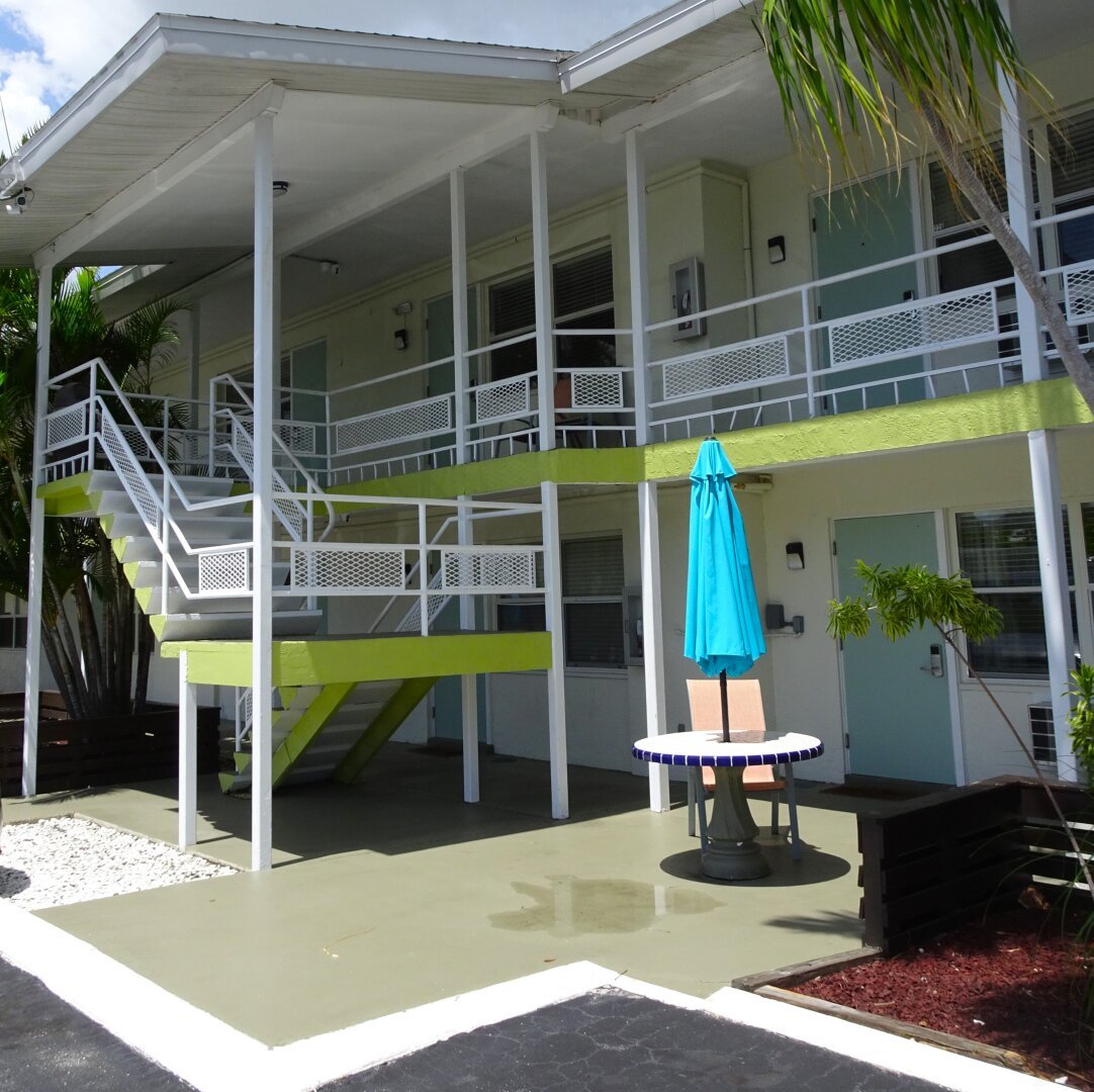 Exterior of a midcentury modern motel. Open concept stairs with white painted metal railings and metal grates. Concrete trim is painted in chartreuse green, doors are painted robin's egg blue. A concrete picnic table accented with cobalt blue tiles and a bright turquoise umbrella sits out front.