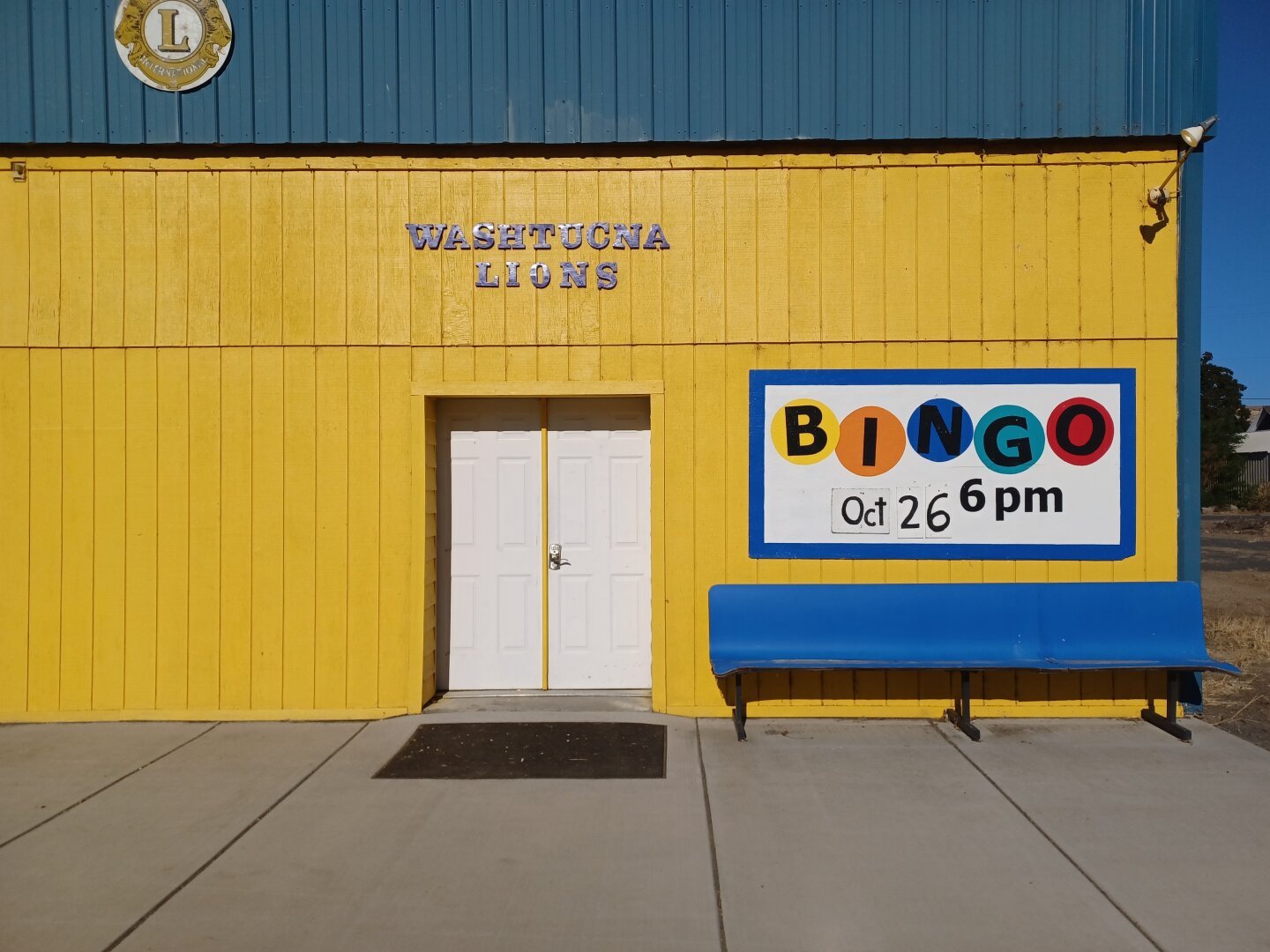 Front of the Washtucna, Washington Lions Hall. The building is painted a very bright yellow. It has a white door and bright blue roofing. A blue bench out front contrasts against the yellow. Over the bench is a brightly colored sign that says 