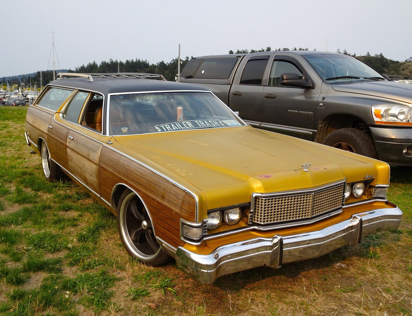 Mid-1970s Mercury Colony Park Wagon, goldenrod-sienna color, with fake wood paneling, in a gravel parking lot. Through the windshield on the dashboard sits a metal sign that says 