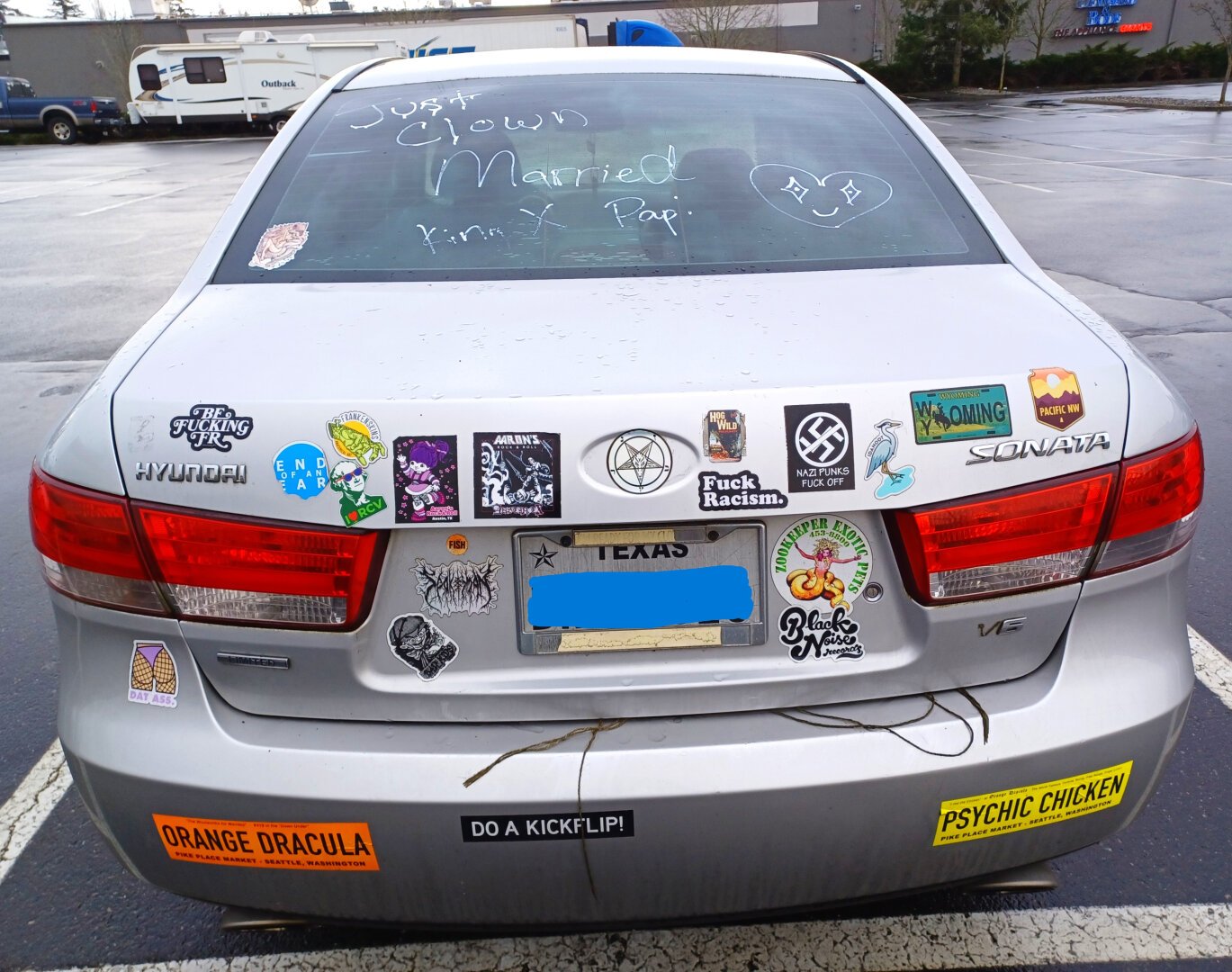 The rear of a silver Hyundai Sonata with the following bumperstickers: Be Fucking FR, I love RCV, Aaron's Rock and Roll Austin TX, Fuck Racism, Nazi Punks Fuck Off, Wyoming, Pacific NW, Dat Ass, Zookeeper Exotic Pets, Black Noise Records, Orange Dracula Pike Place Market Seattle Washington, Psychic Chicken Pike Place Market Seattle Washington, Do a Kick Flip. The back window has some white handwriting that says 