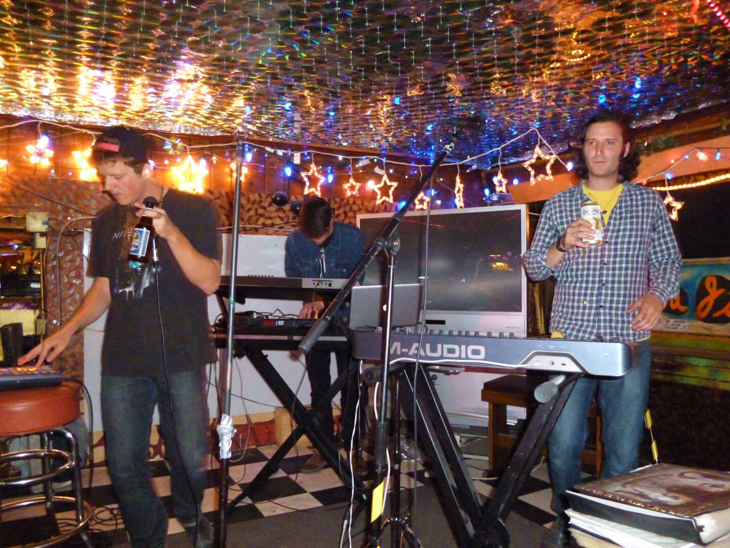 Three members of TV Girl inside the Java Jive. They each have a synthesizer keyboard. Two of the band members are holding beers. The low ceiling above the stage is shiny and twinkling with small lights.