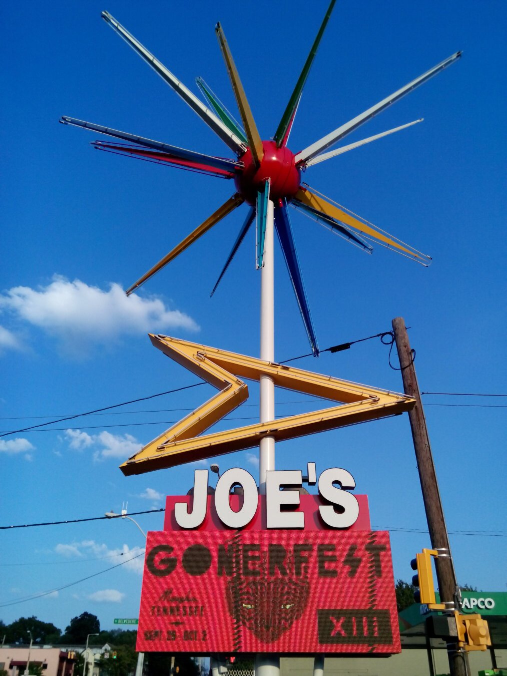 Roto-sphere neon sign against a blue sky. The sign has a large central sphere with large neon spikes radiating from it. Below the sphere is a large yellow neon arrow pointing right, and below that is a digital readerboard that says 